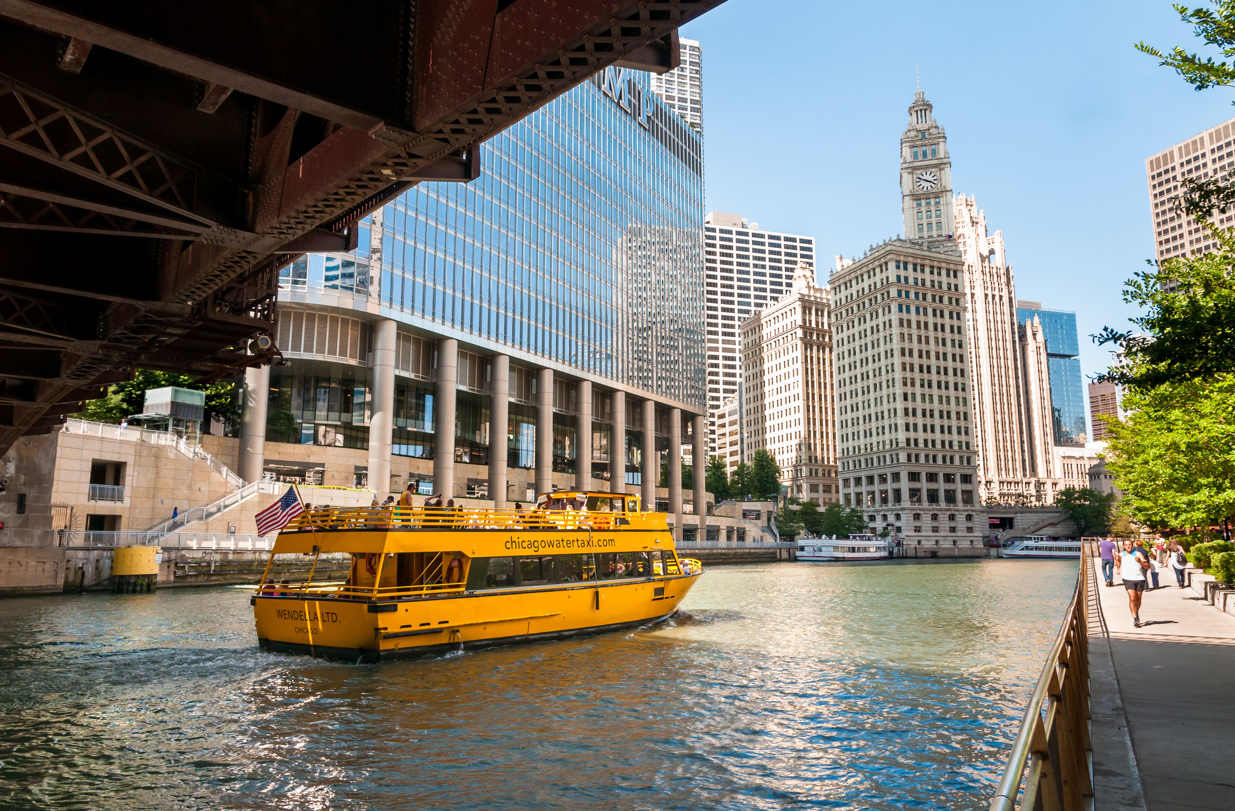 A bright yellow, two-deck boat passes below a bridge on an urban river. A glassy blue skyscraper is visible on the far bank and a pedestrian filled riverwalk on the near side.