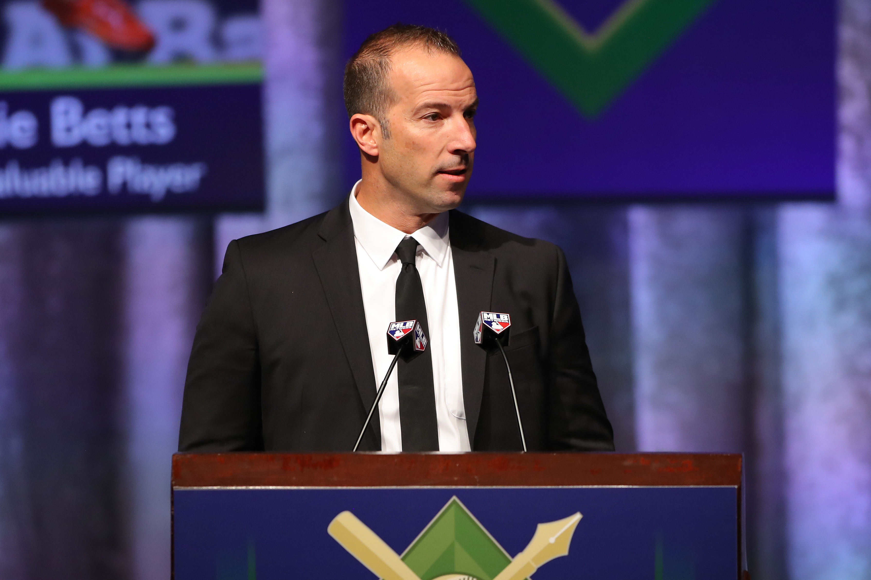 Billy Eppler wearing a black suit and black tie but no sunglasses standing at a podium with two microphones.