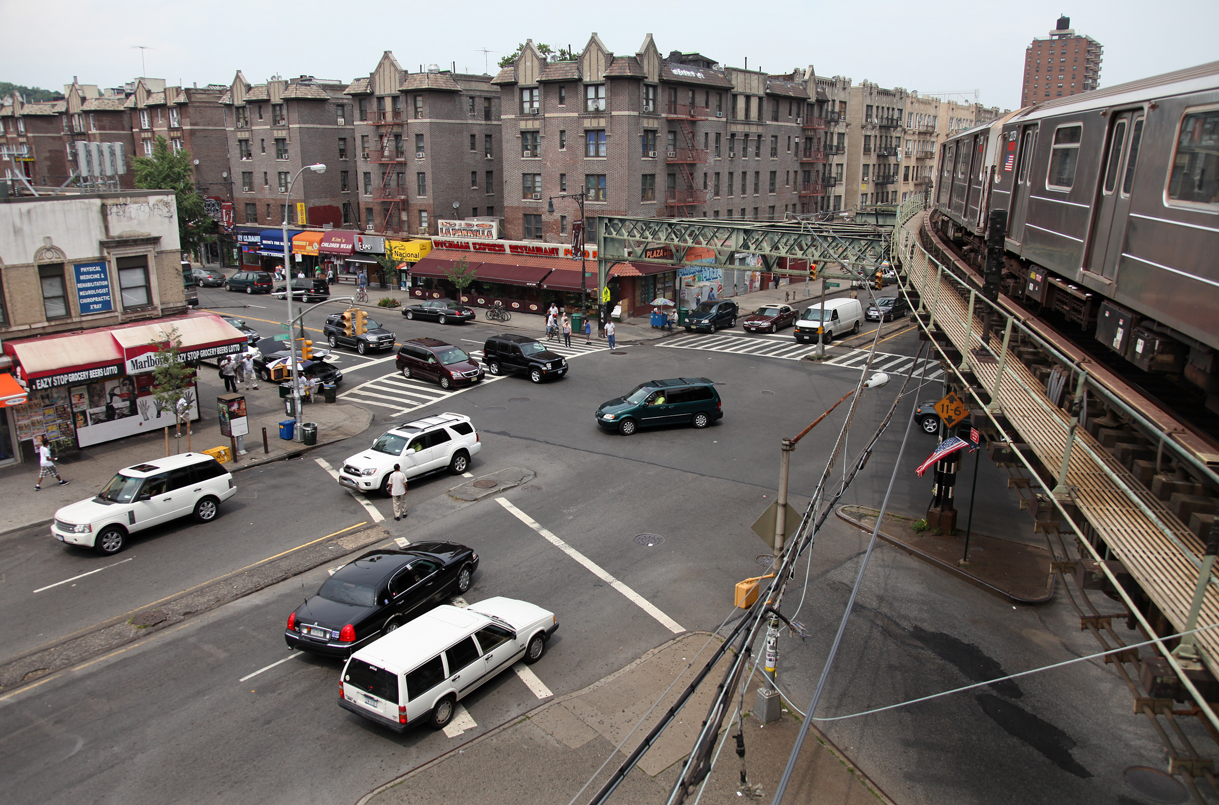 A busy intersection in Inwood, Manhattan is shown with cars zipping through. A train passes on the above ground subway over the intersection. A row of apartment buildings are in the background.