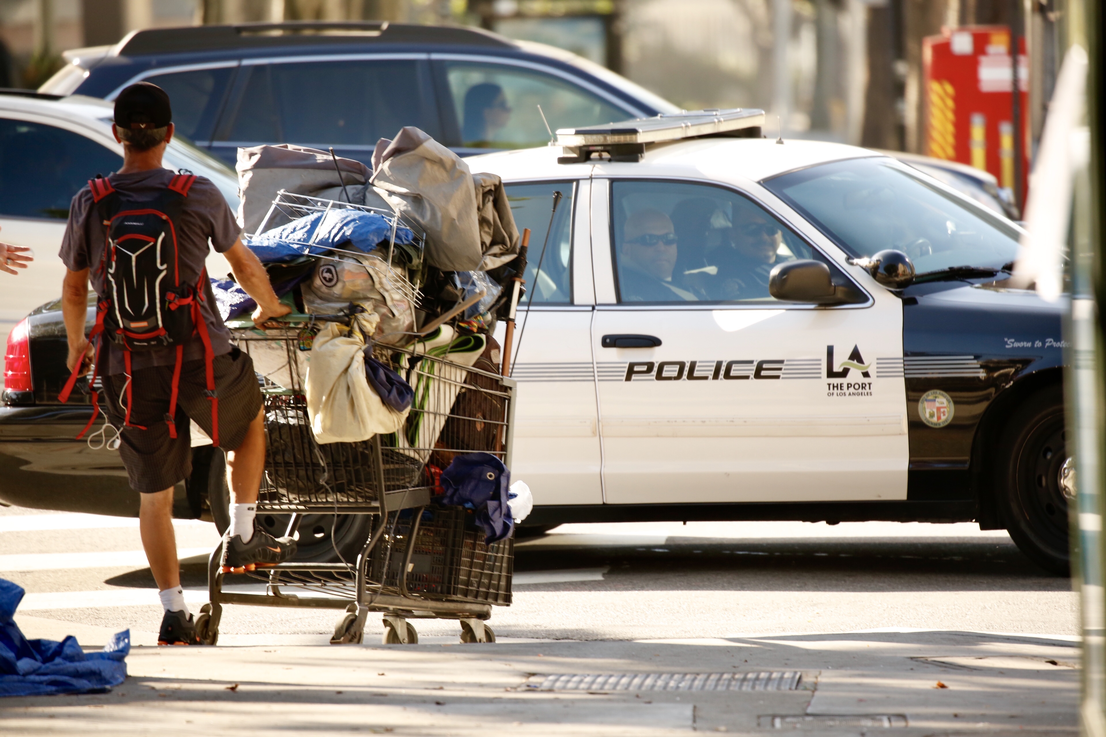 A ban with a backpack pushes a cart full of belongings down a street. A police car is present in the background.