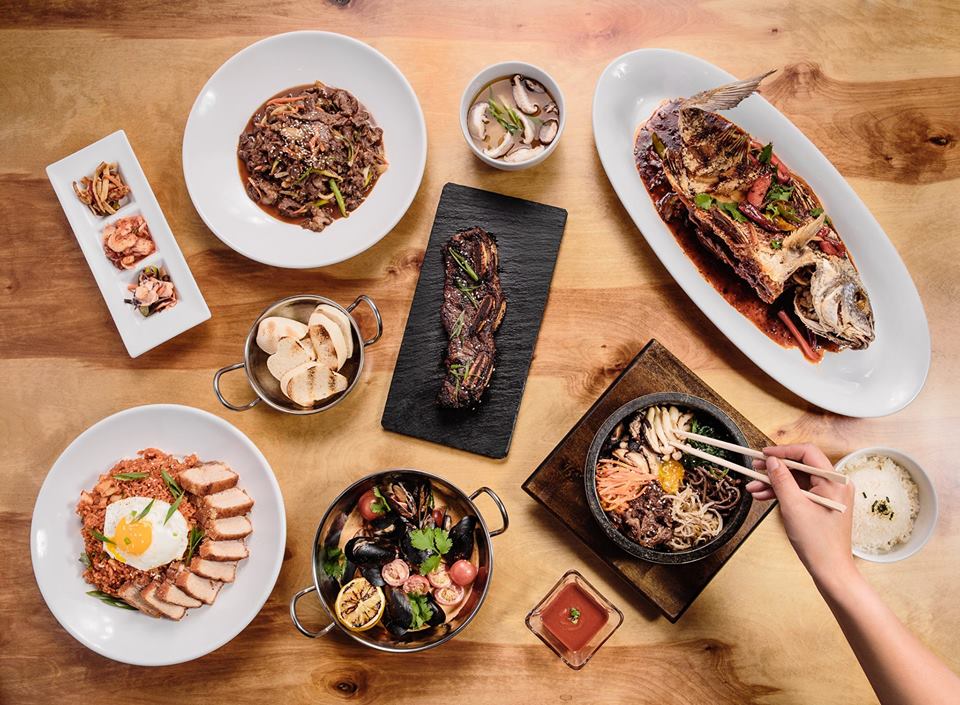 An array of dishes and trays with food from Jenna’s Asian Kitchen, from rice and sliced meats to toasted breads to a whole fish, to someone picking up food with chopsticks