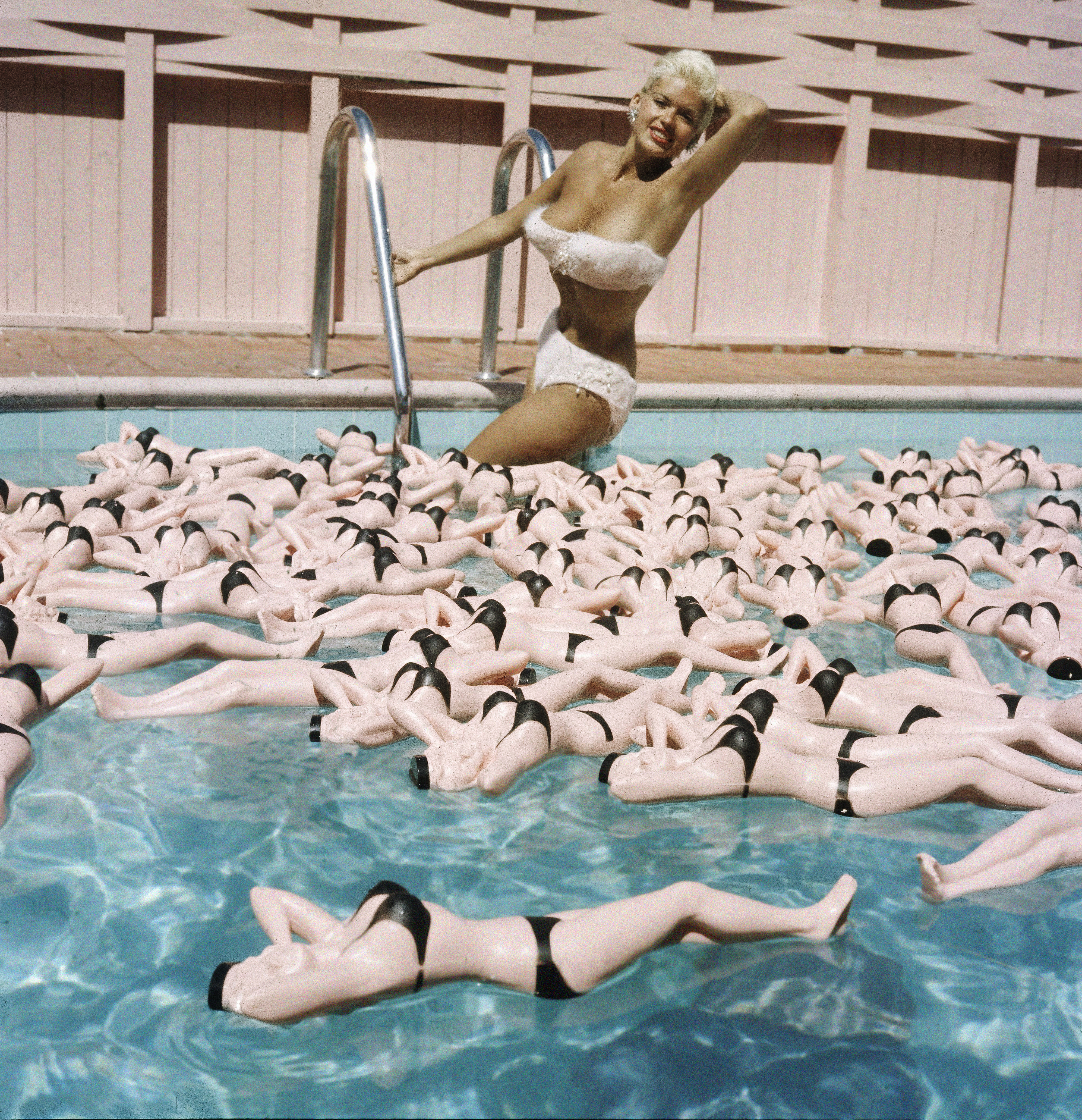 Jayne Mansfield, with a tan and platinum blond hair, poses in a white bikini at the edge of a swimming pool filled with bottles shaped like bikini-clad versions of herself.