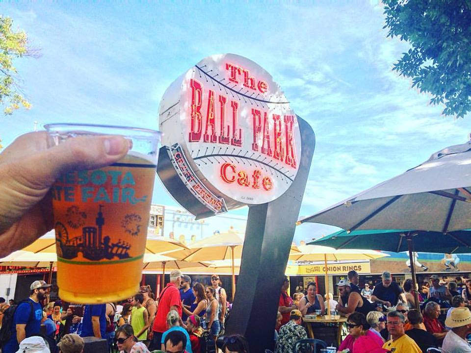 A hand holds up a beer next to the baseball shaped neon sign for the Ball Park Cafe on a busy fair day