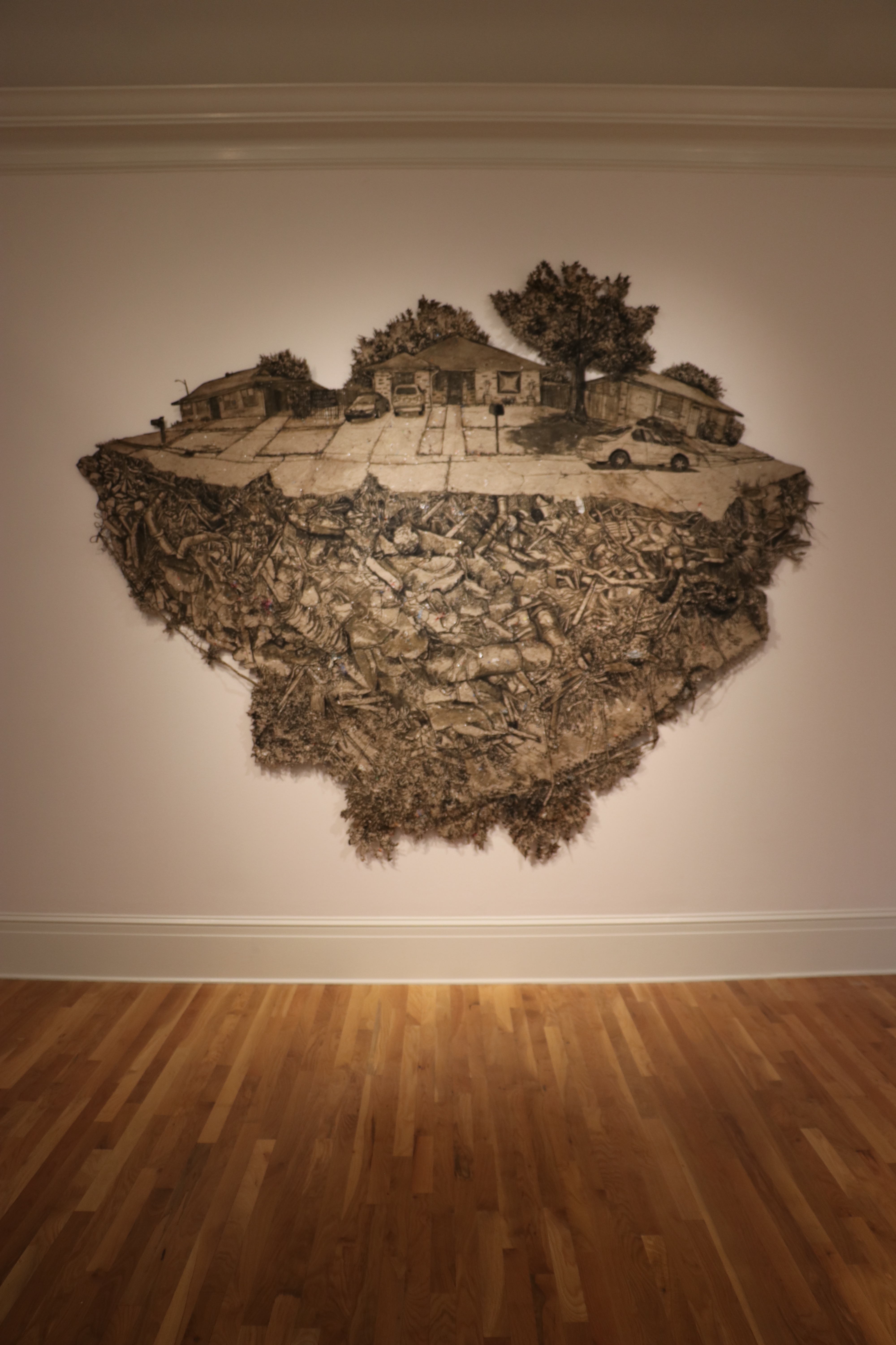 A handmade, irregularly sized piece of paper printed with images of a neighborhood atop a landfill hangs on a museum wall