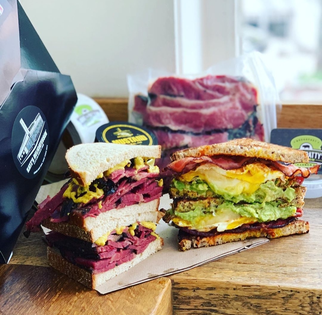 Two halves of a pastrami sandwich sit stacked next to two halves of an eggy breakfast sandwich
