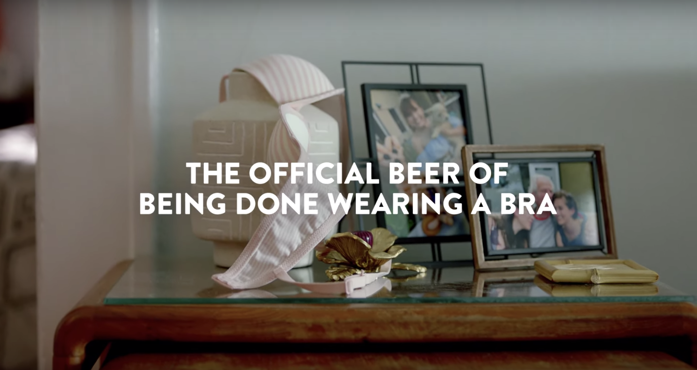 A woman’s discarded bra lays slung over framed photos on a glass side table with the words “The official beer of being done wearing a bra.”