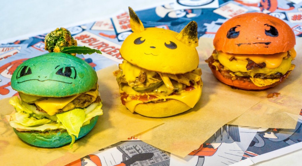 Burgers with a green bun, a yellow bun, and a red bun sit on napkins on a table