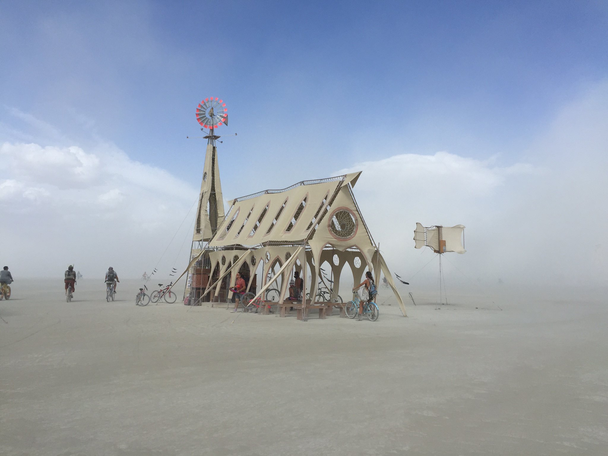 A wooden makeshift church temple with a red windmill on top in the dusty desert.