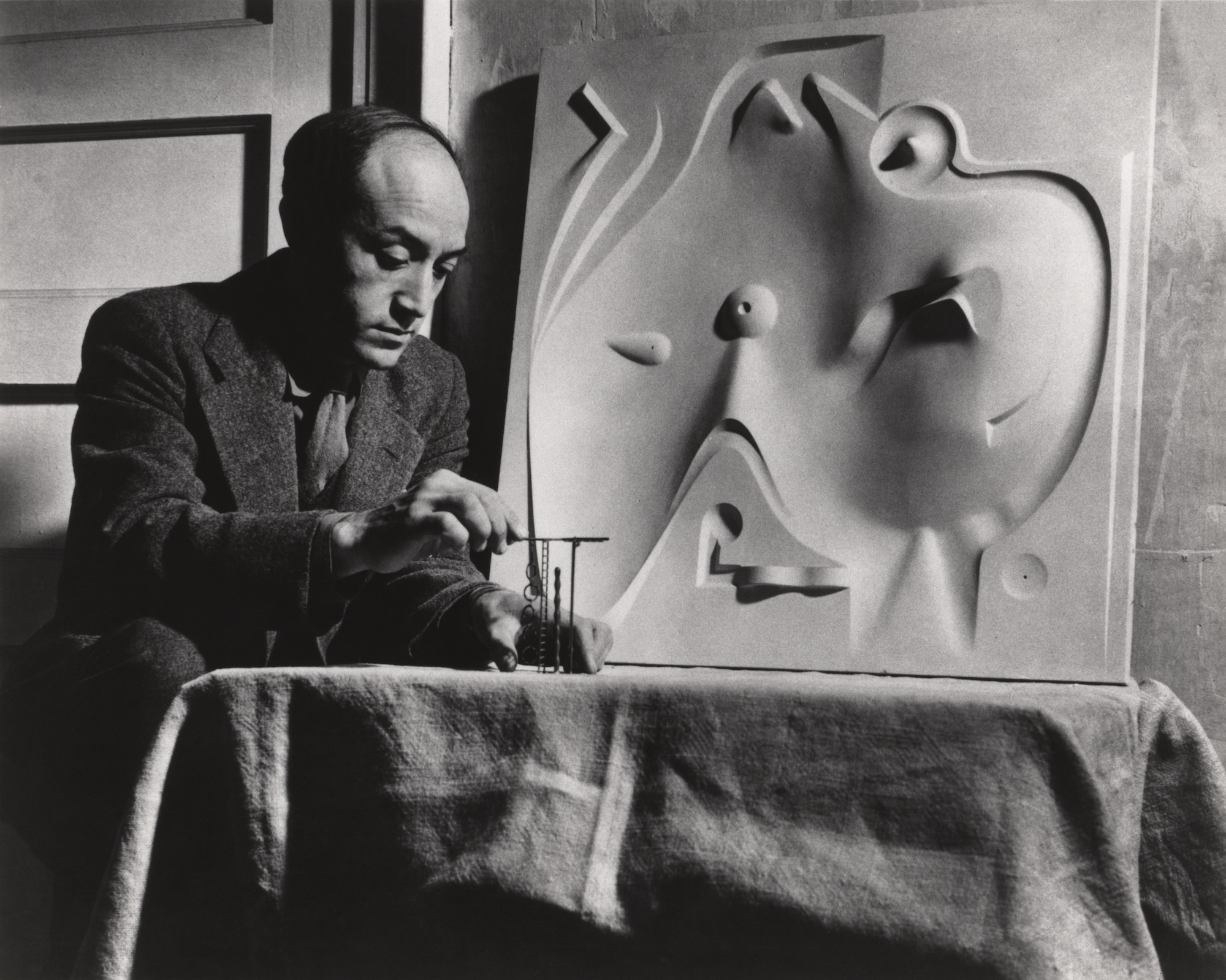 A man sits in front of a bas-relief plaster sculpture of an undulating playscape while looking at tiny versions of abstract structures