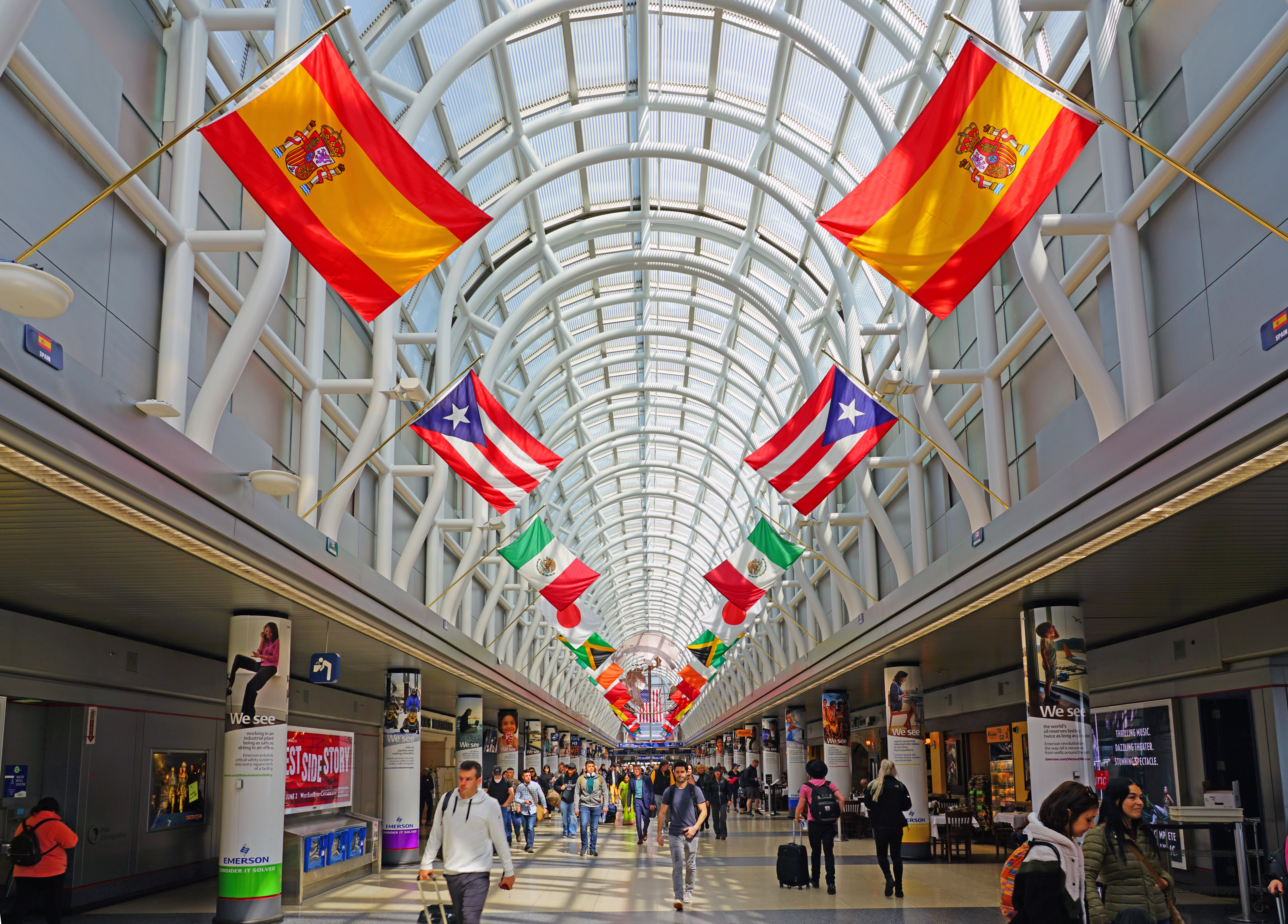 A glass-dome massive hallway at an airport with gates in the distances. International flags decorate columns up top near the skylight. 