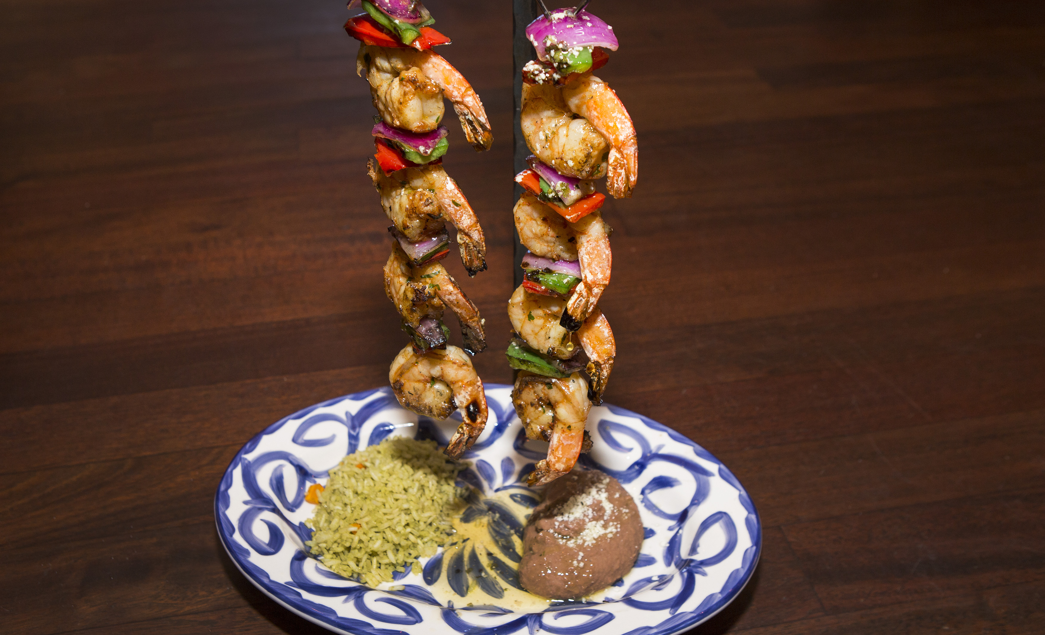 Two skewers of shrimp, onion, red, and green peppers hang on a metal stand over a blue and white plate that contains Mexican rice and refried beans.