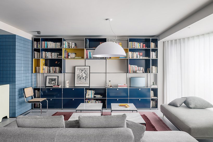 Inside a modern apartment living room, a wall of blue shelving holds books and artworks. Light gray sofa and lounge surround a set of four white marble top coffee tables. 