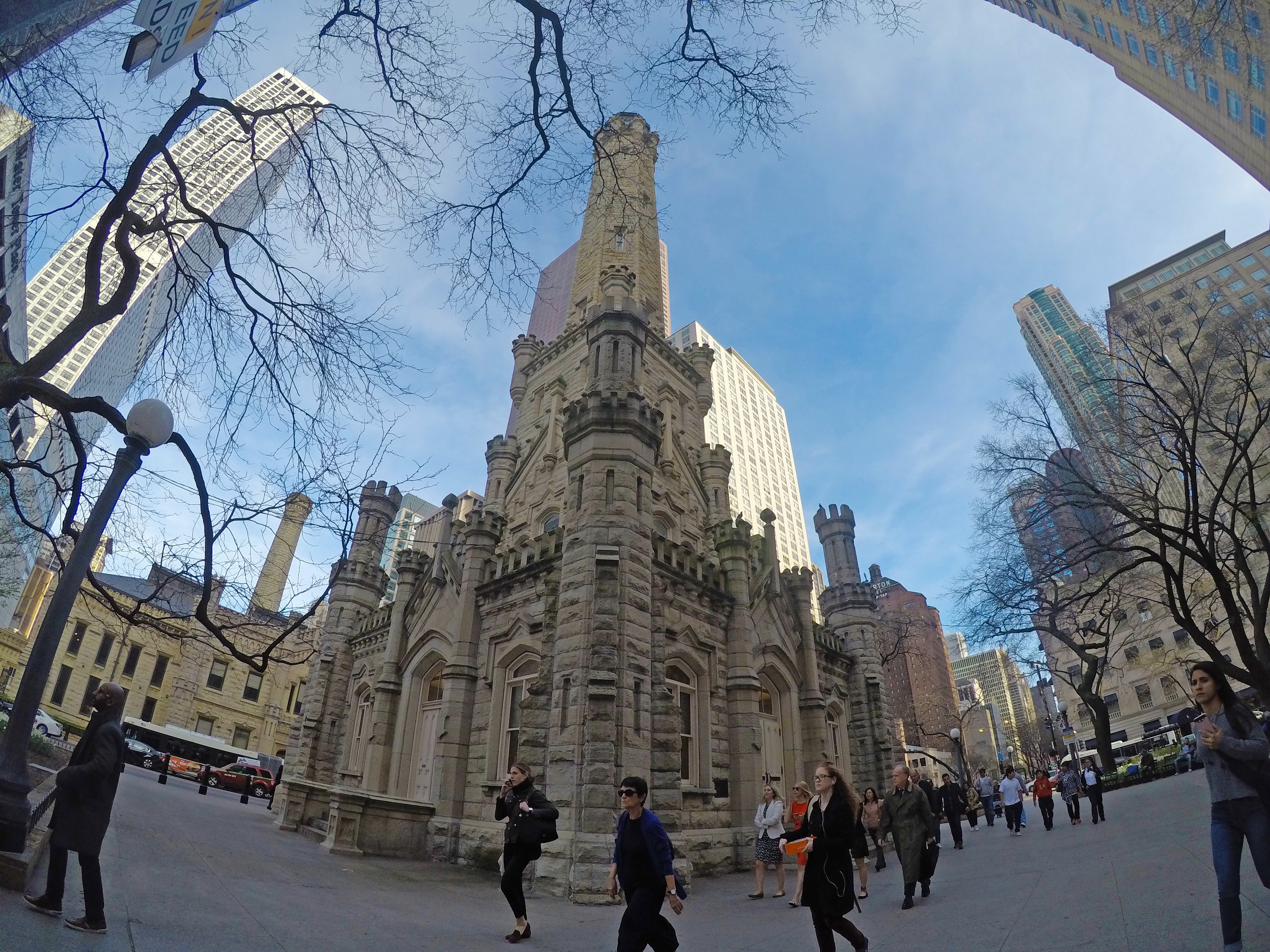 With a fish eye lens, a photo of the small, square stone building with a tall spire. People walk past the structure on a busy downtown block. 
