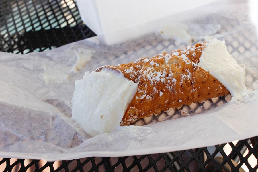 A cannolo from Maria’s Pastry Shop in Boston’s North End, garnished with powdered sugar and stuffed with ricotta, sits on tissue paper on an outdoor table.