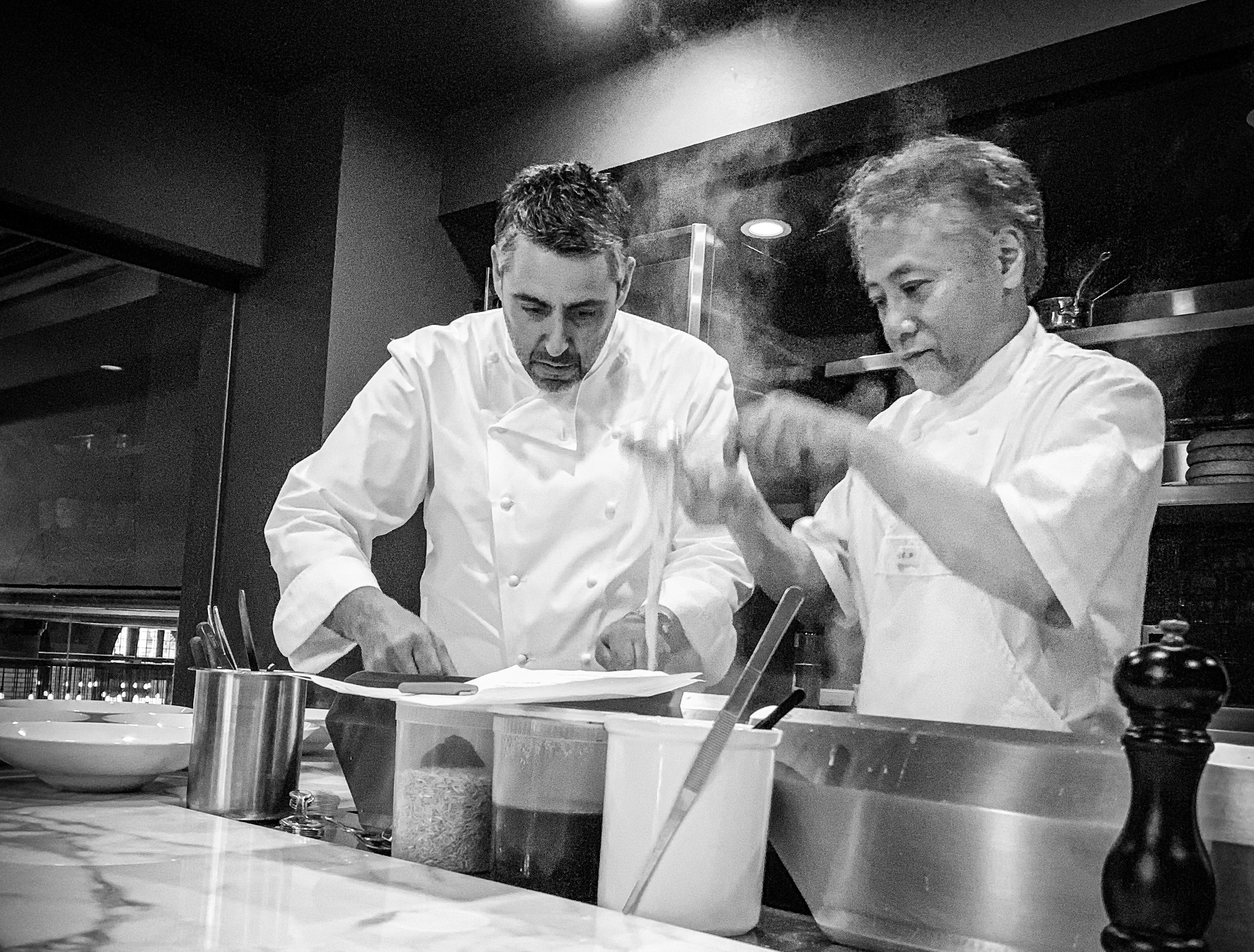 A black and white photo of chef Luciano DelSignore and chef Takashi Yagihashi standing in a kitchen in front of a steaming pot and a marble counter wearing white chefs coats.