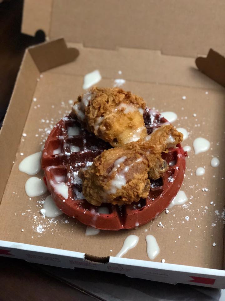 A bright red waffle — red velvet flavor — sits in an open pizza box and is topped with a couple pieces of fried chicken and a spattering of icing