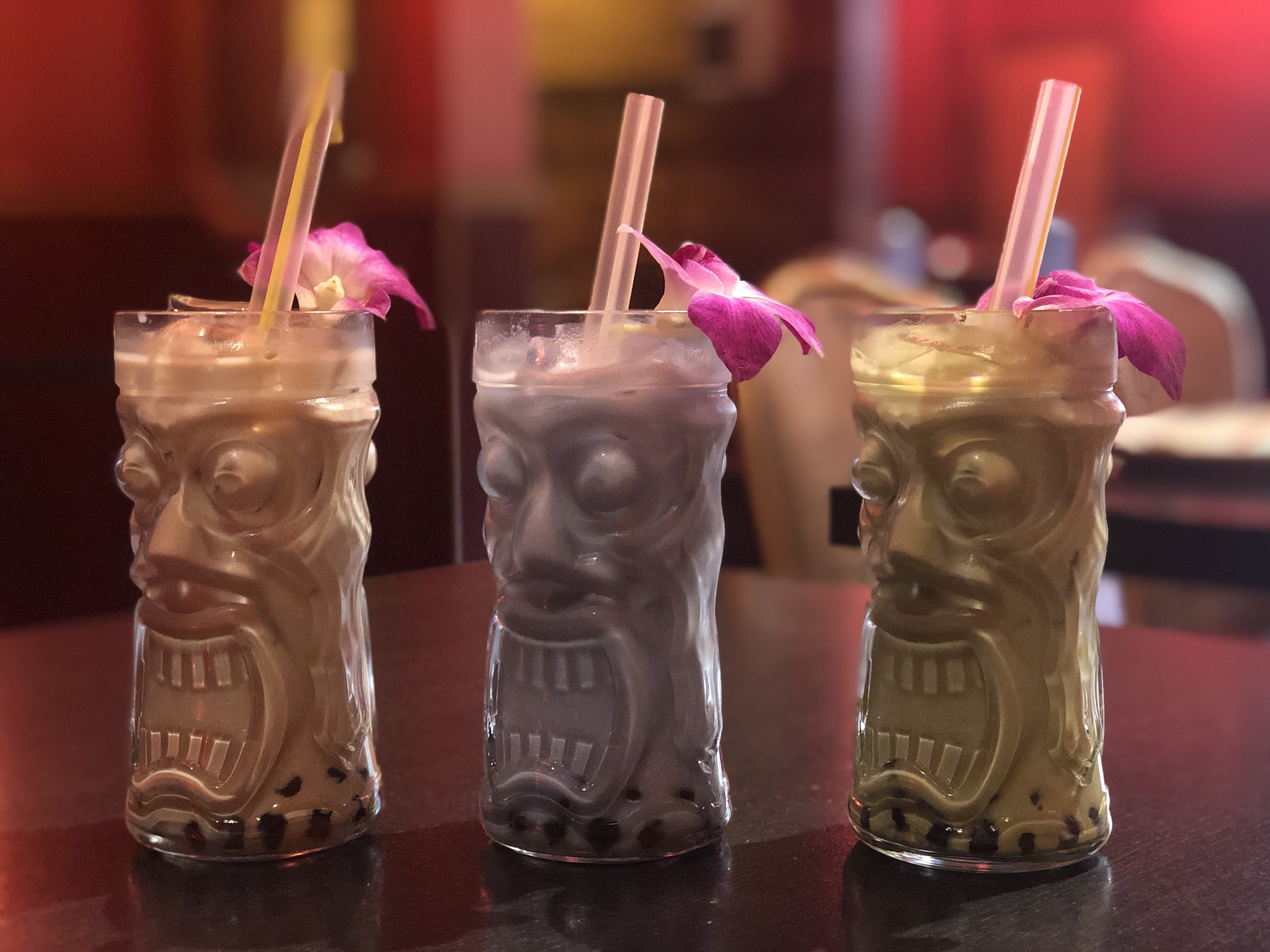 A row of three clear glass Tiki mugs with boozy boba cocktails in three colors. Each mug is garnished with an orchid and colored straw.