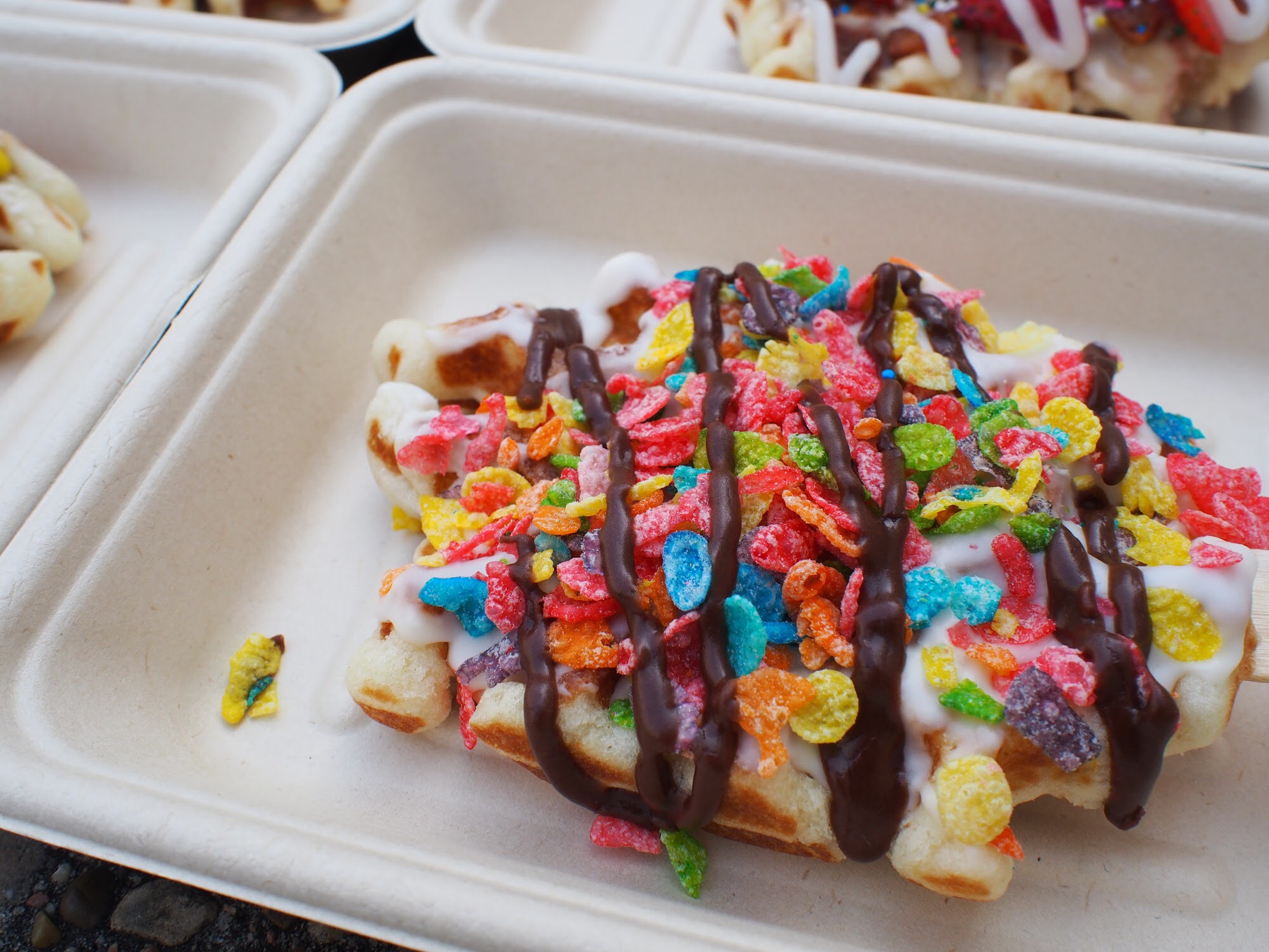 A Belgian waffle topped with colorful Fruity Pebbles cereal and white sauce and a chocolate drizzle