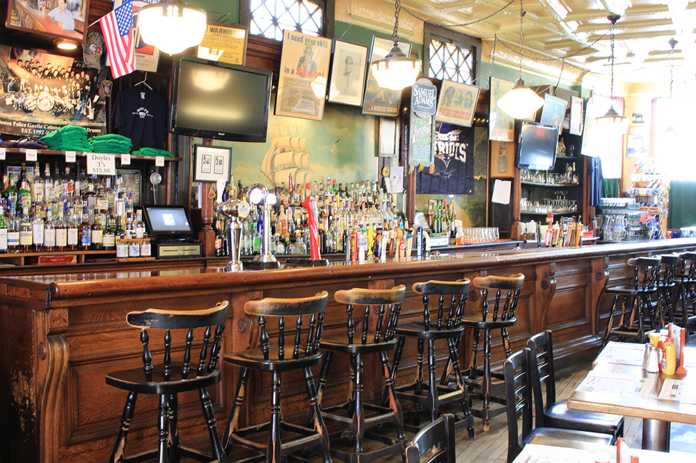 The bar at Doyle’s Cafe, with high chairs lined up in front of a wooden bar, sunlight streaming in, and a television, American flag, and other decor on the green wall