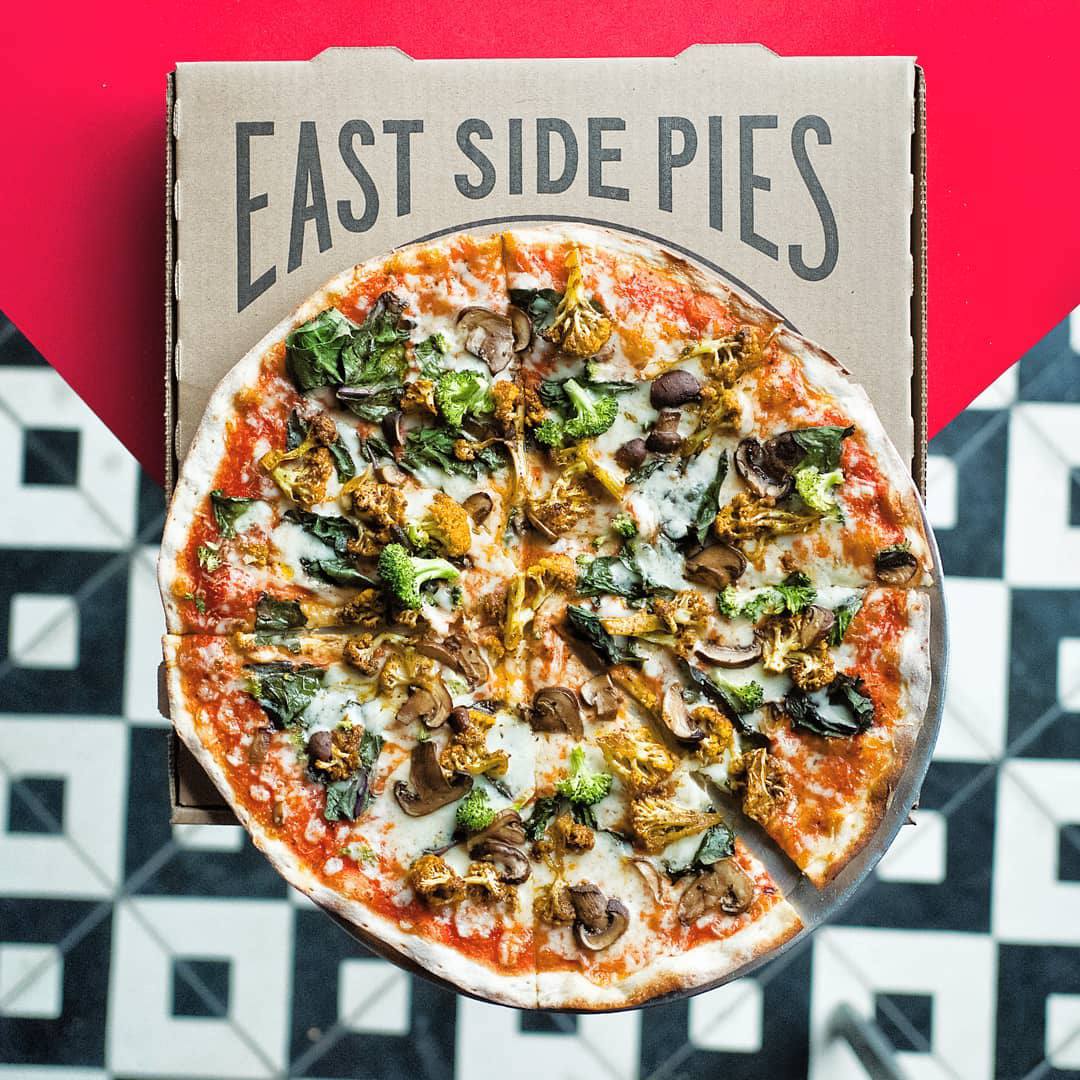 A pizza pie topped with cheese, sauces, and vegetables on top of a pizza box with the words East Side Pies on it