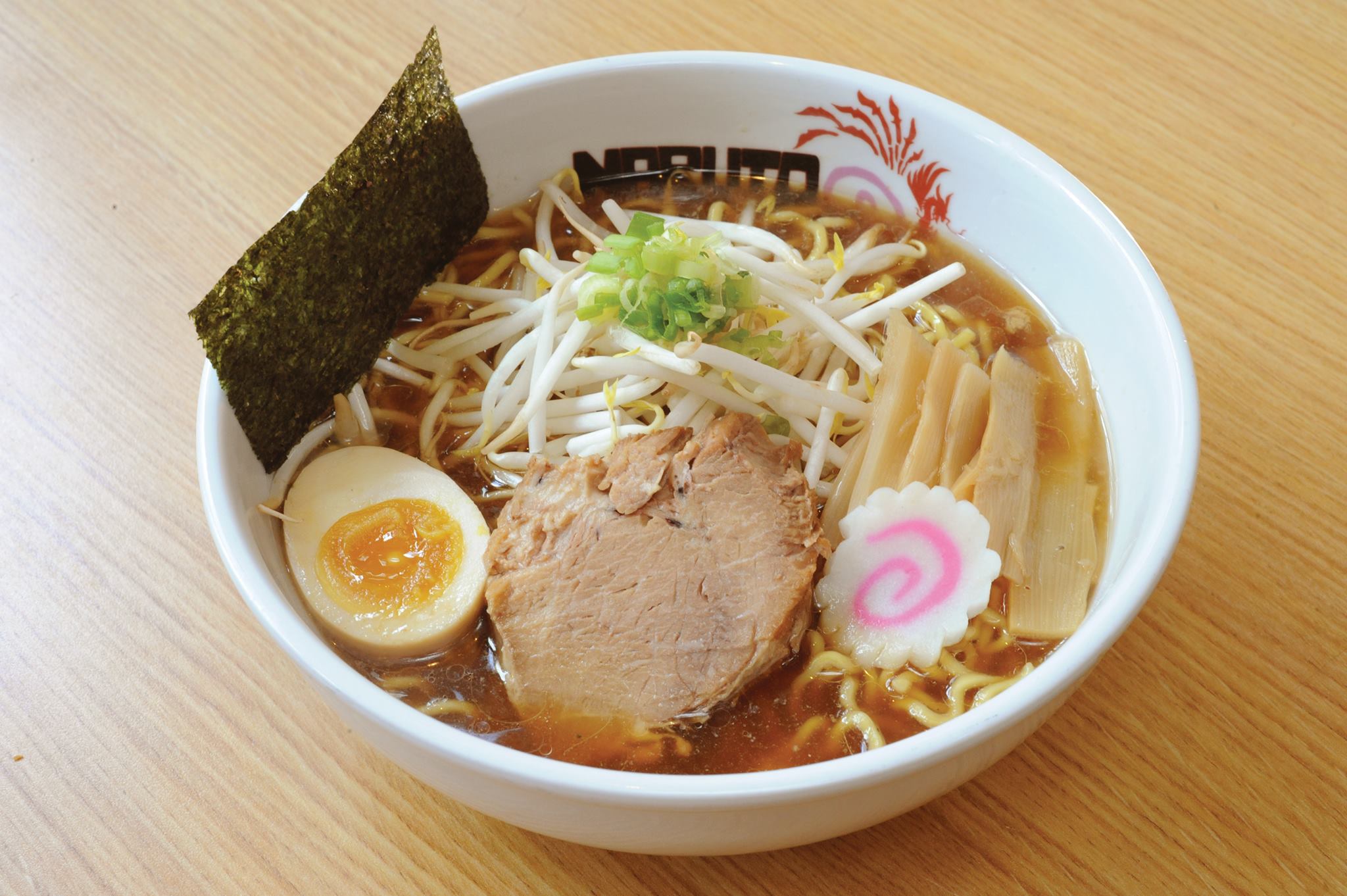 A bowl of ramen with soup, noodles, bean rpouts, greens, a slice of pork, a half-sliced hard-boiled egg, a fish cake with a pink swirl, and a slice of seaweed