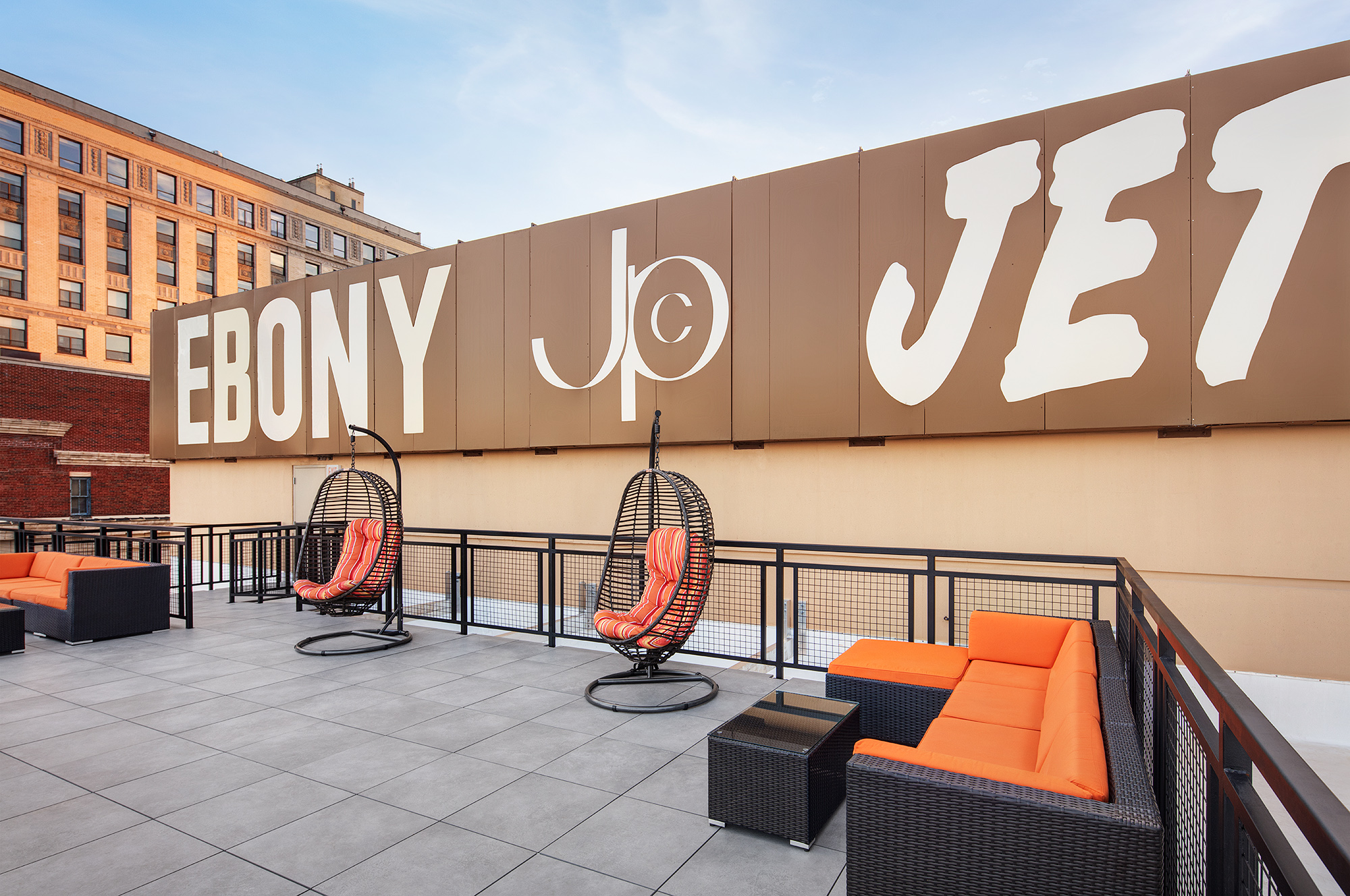 A rooftop with two swinging chairs and a few lounge couches sit in front of a large brown and white sign that displays the historic “Ebony, JPC, Jet” text.