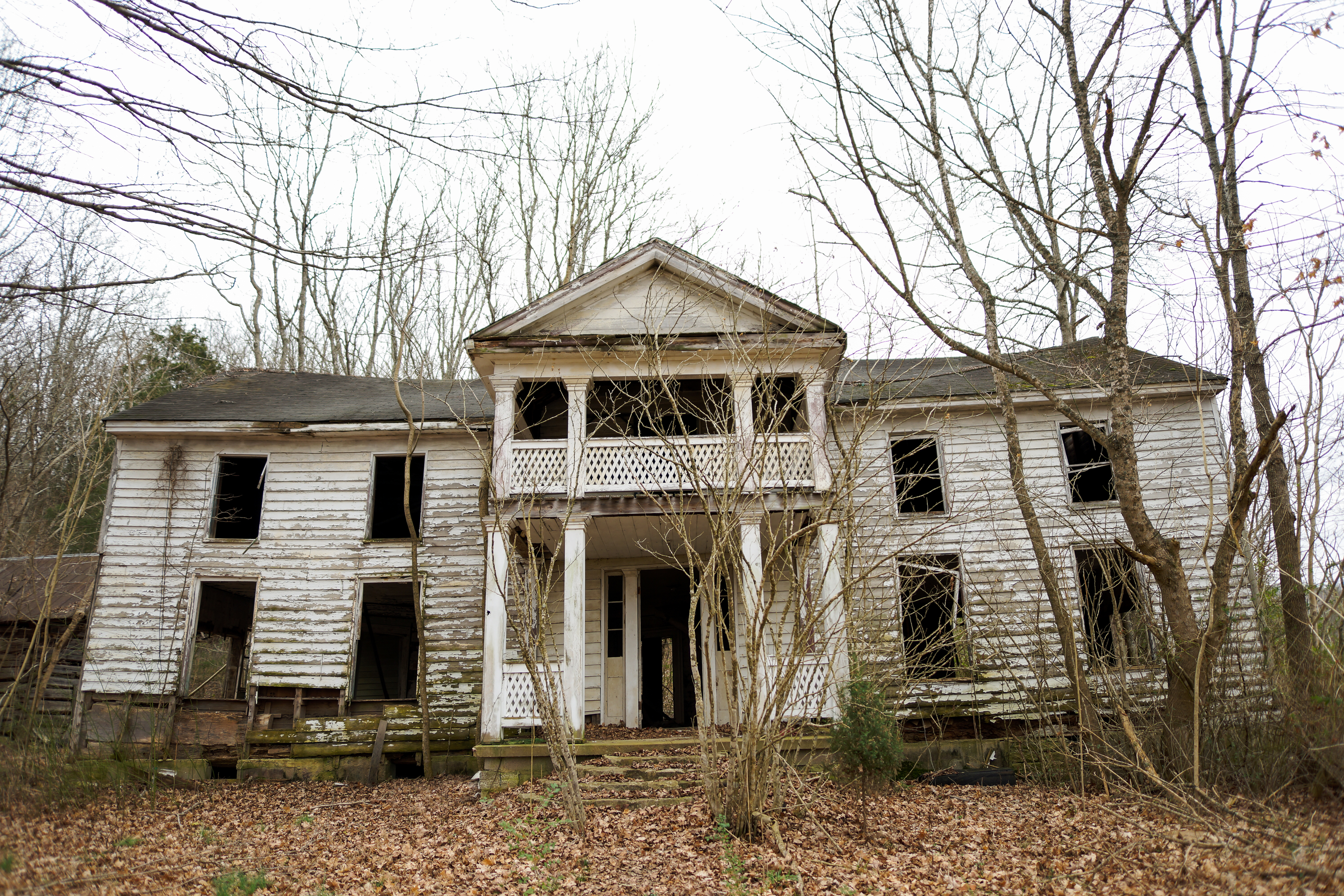 A dilapidated colonial-style white house in the colonial style sits in a woodland setting with lots of dead trees. 