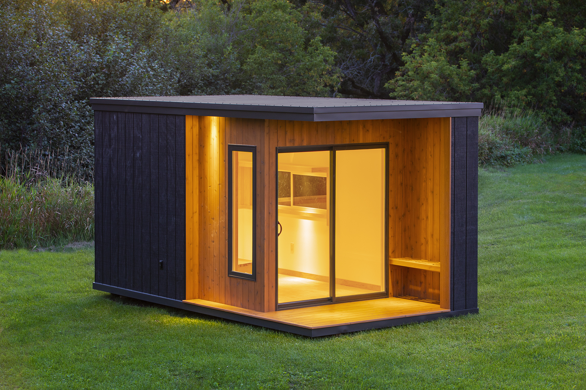 A small boxy dwelling sits on a grassy plot. It is clad in black and natural wood, with a small covered porch and sliding glass doors to the inside.