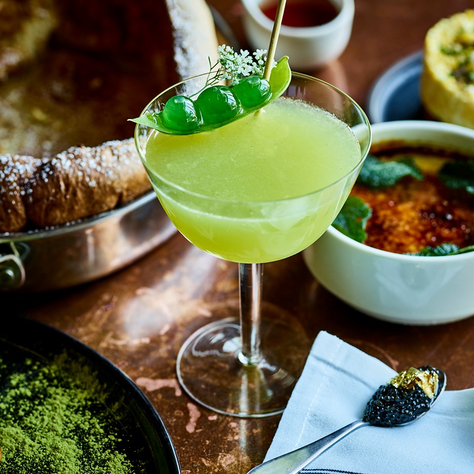 A green cocktail in a coup with peas on top surrounded by creme brulee, a spoon with black caviar and gold leaf, and a Dutch baby pancake.