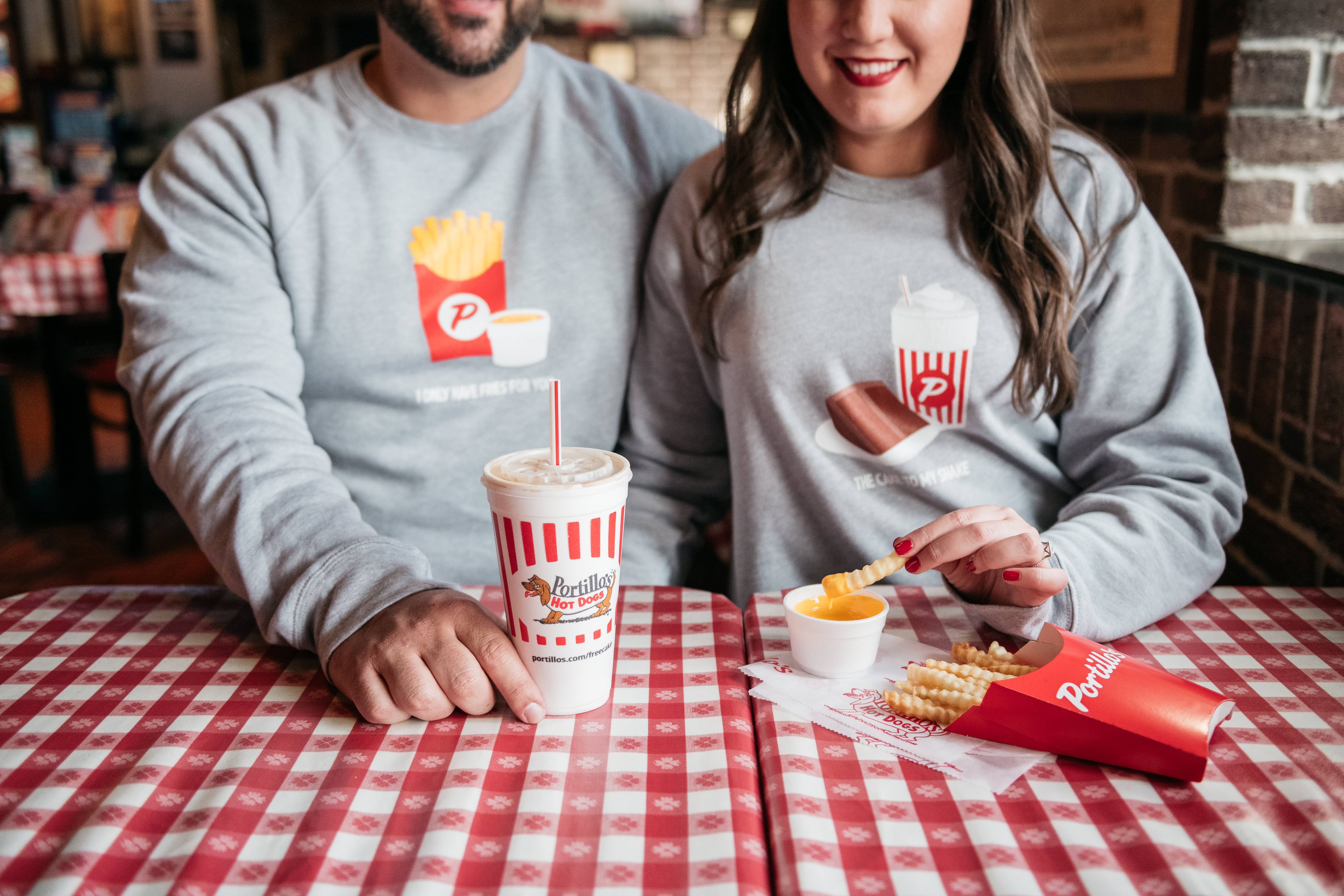 A white man and white woman sit next to each other at a table covered with a checkered cloth. They hold a container of fries and a soda cup.