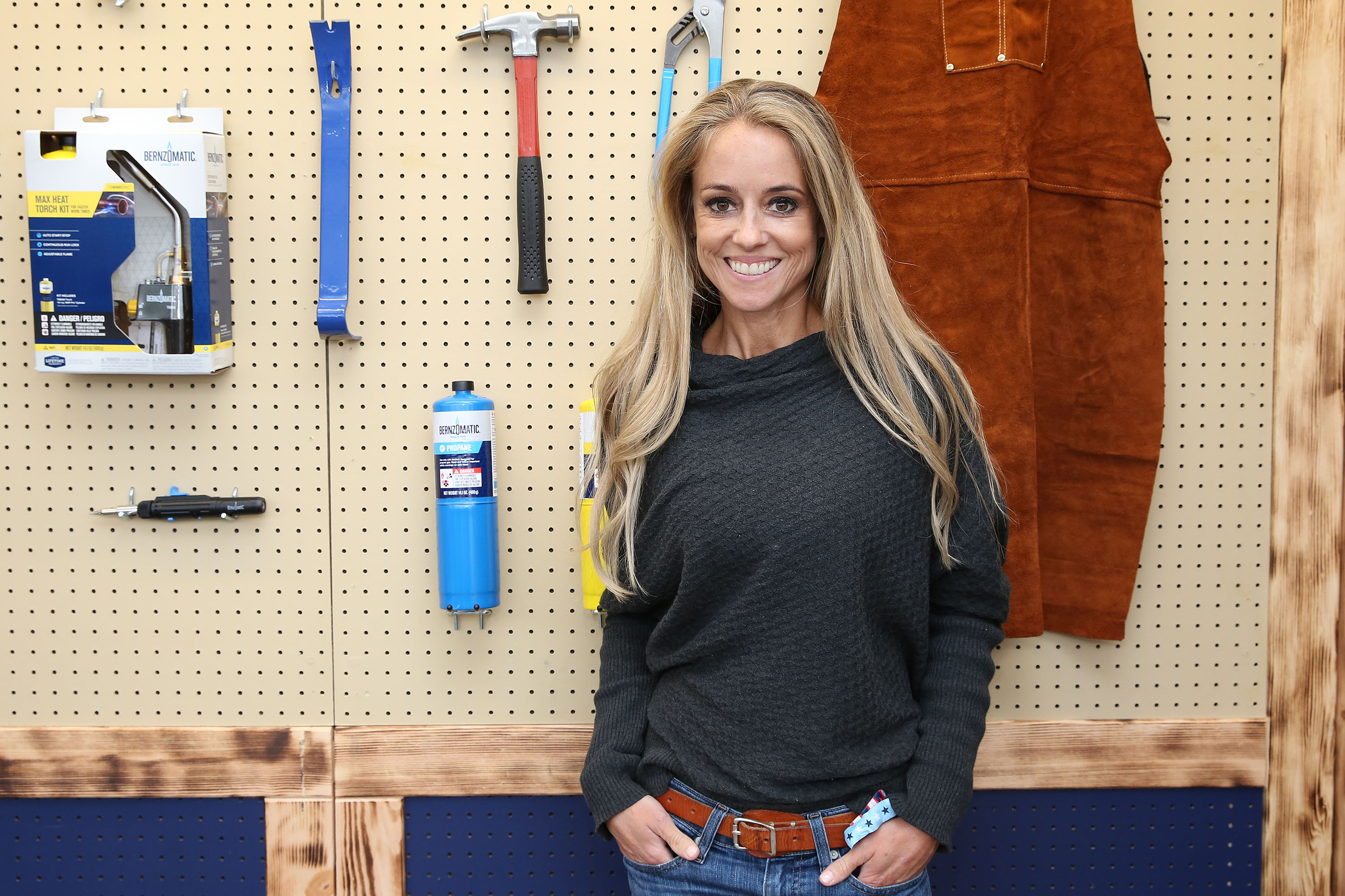 A woman in a black shirt and jeans stands in front of a wall of hanging carpentry tools.