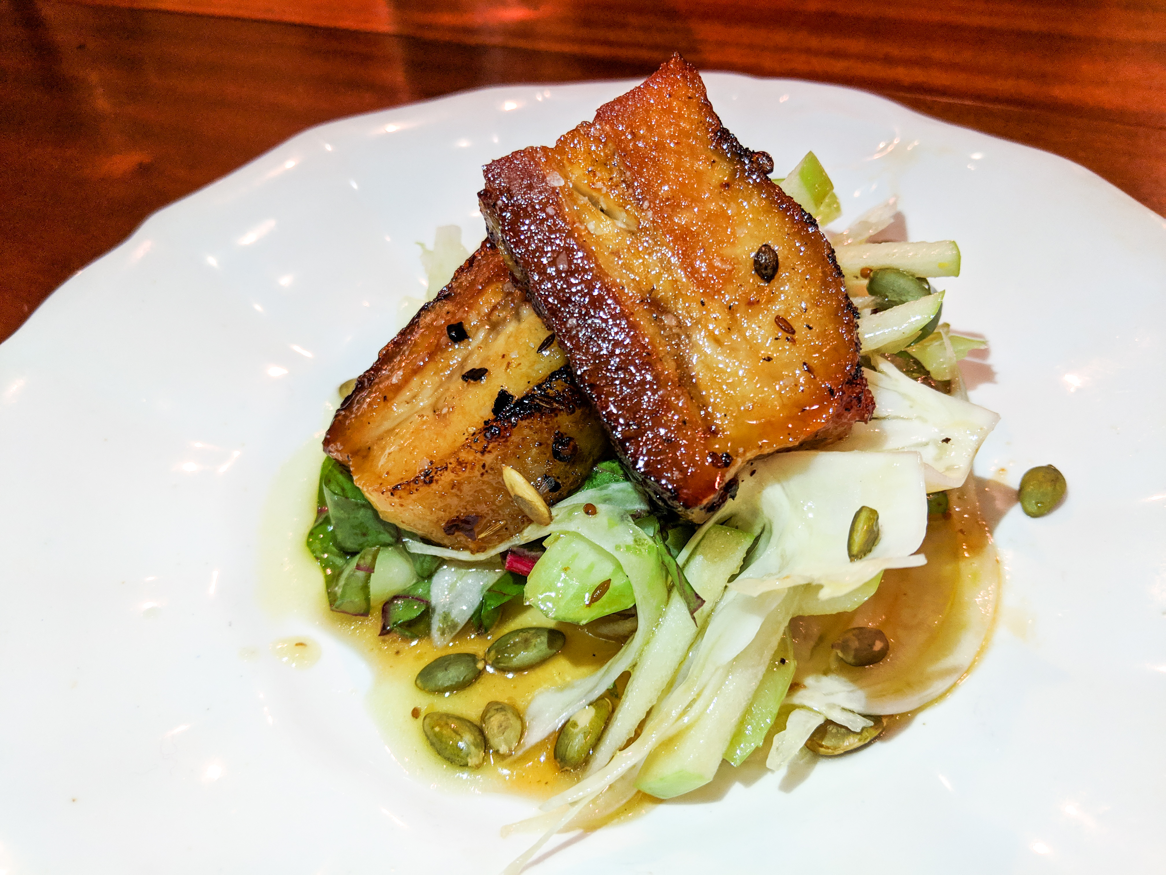 Two slabs of pork belly sit on a white plate with greens and other accoutrements. The plate is on a glossy wood bar.