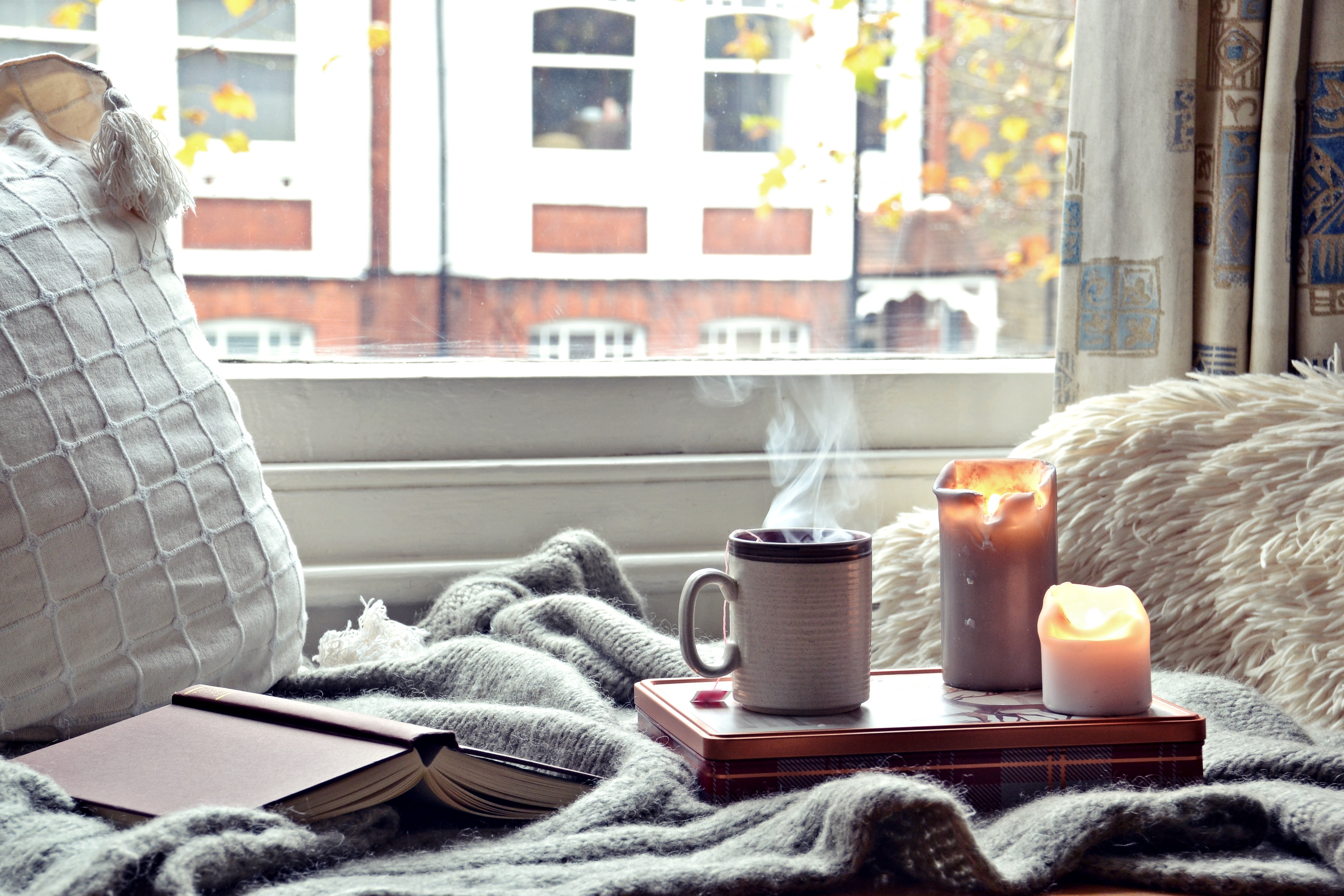 Cozy pillows and blankets sit adjacent to a window that looks out onto an urban street. A book is overturned on the blanket and a tray holds two lit candles and a steaming mug of liquid.