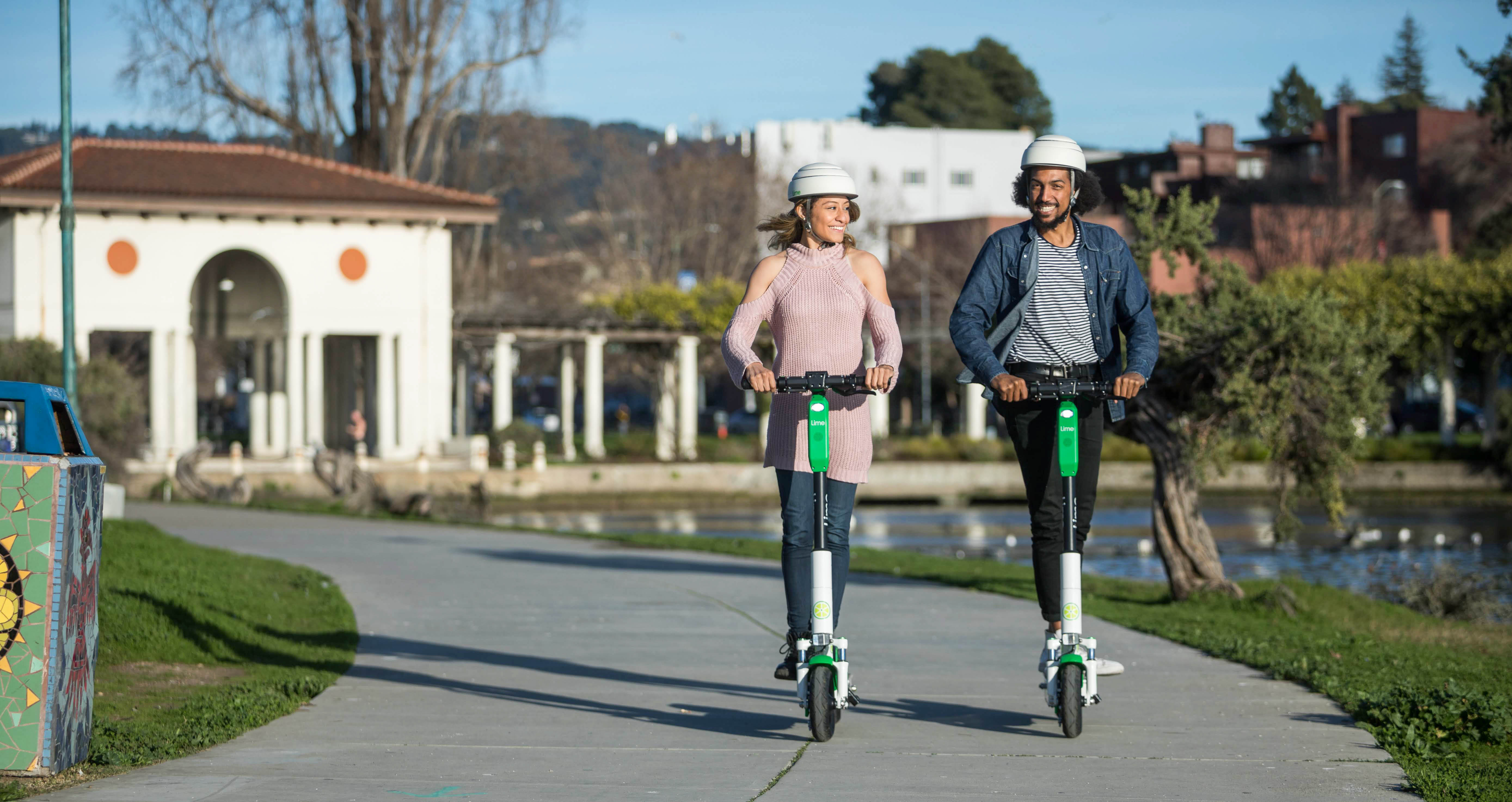 A man and woman, both wearing helmets, each riding their own electronic scooter.