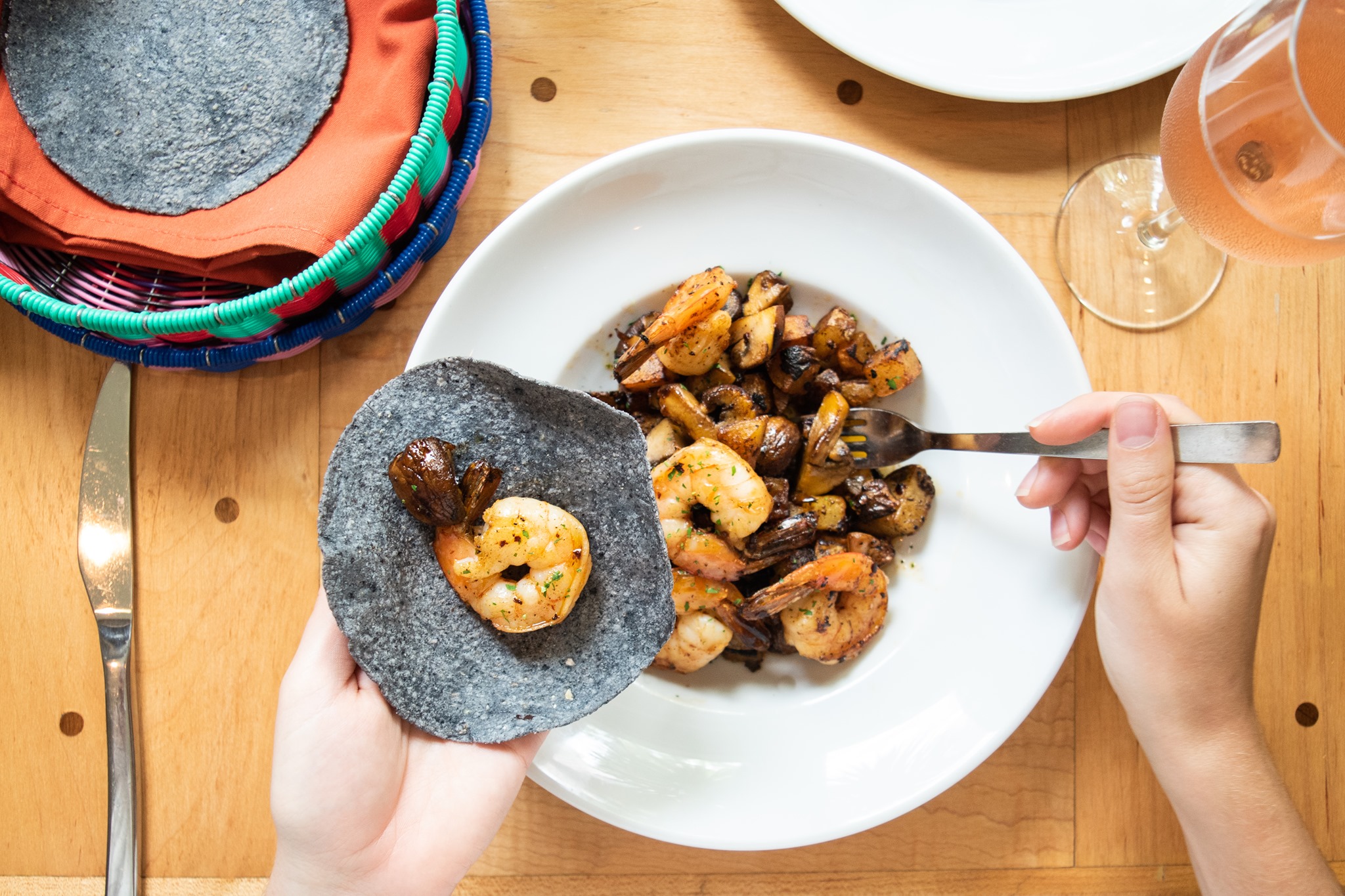 A person holding up a blue tortilla with a shrimp on it with one hand and spooning a bowl of shrimp with the other