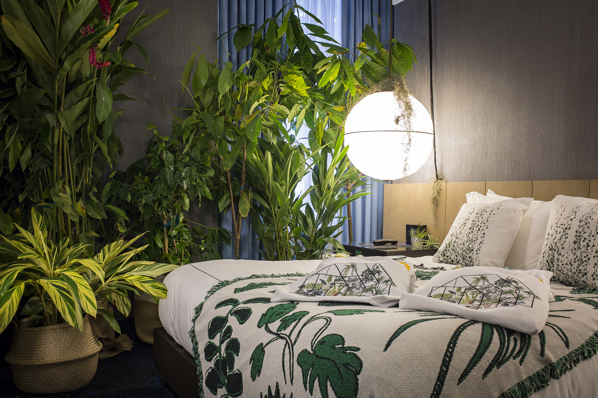 A room with leafy plants, a globe light and bed with embroidered blanket.