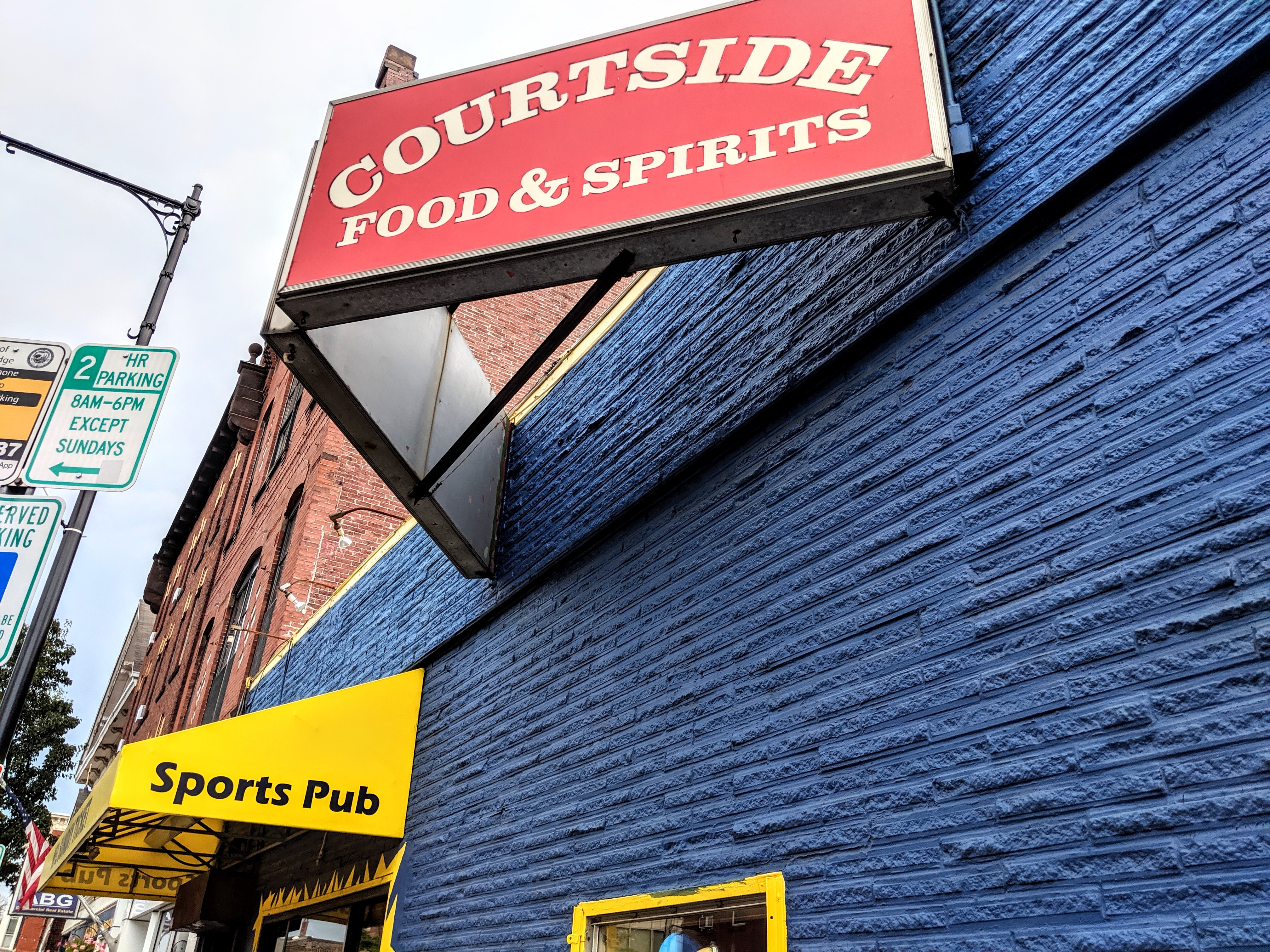 A blue-painted brick building with yellow awnings and a red sign that reads Courtside Food &amp; Spirits