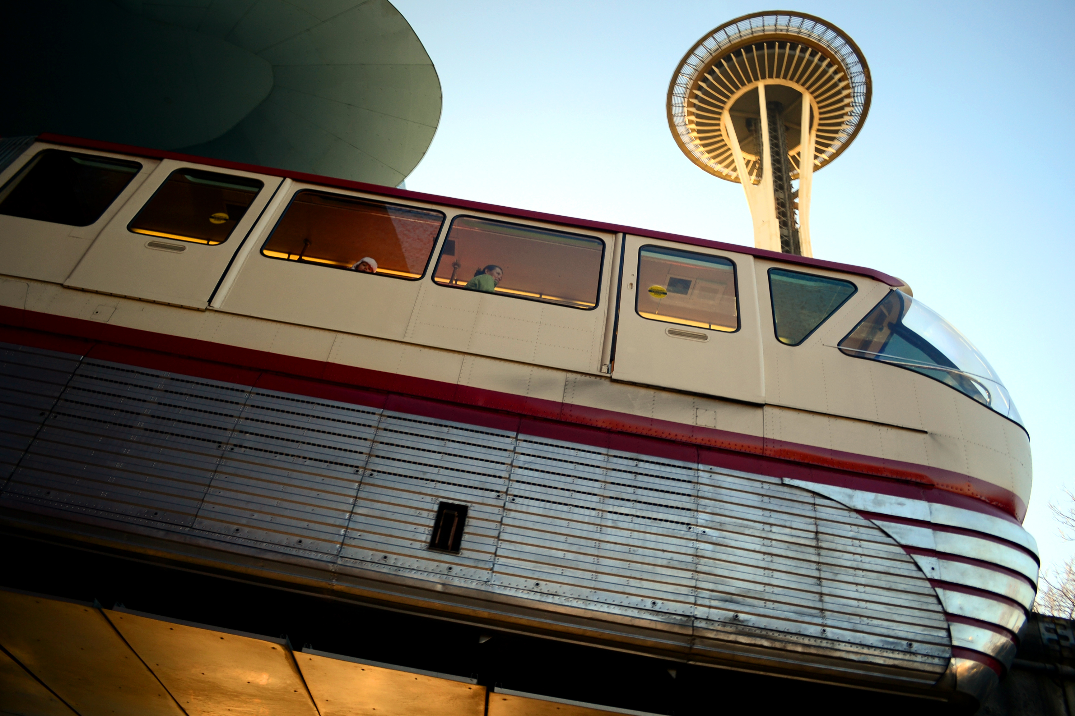 A monorail train exits a tunnel under a blue sky and a tall building with a saucer on top (the Space Needle).