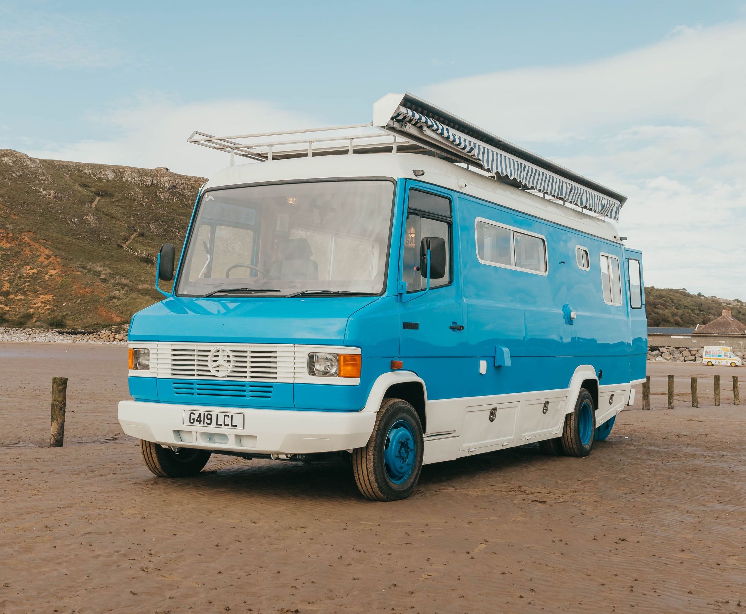 A blue camper van with white trim, large windows, an awning, and a white roof rack. The camper sits on a sandy beach.