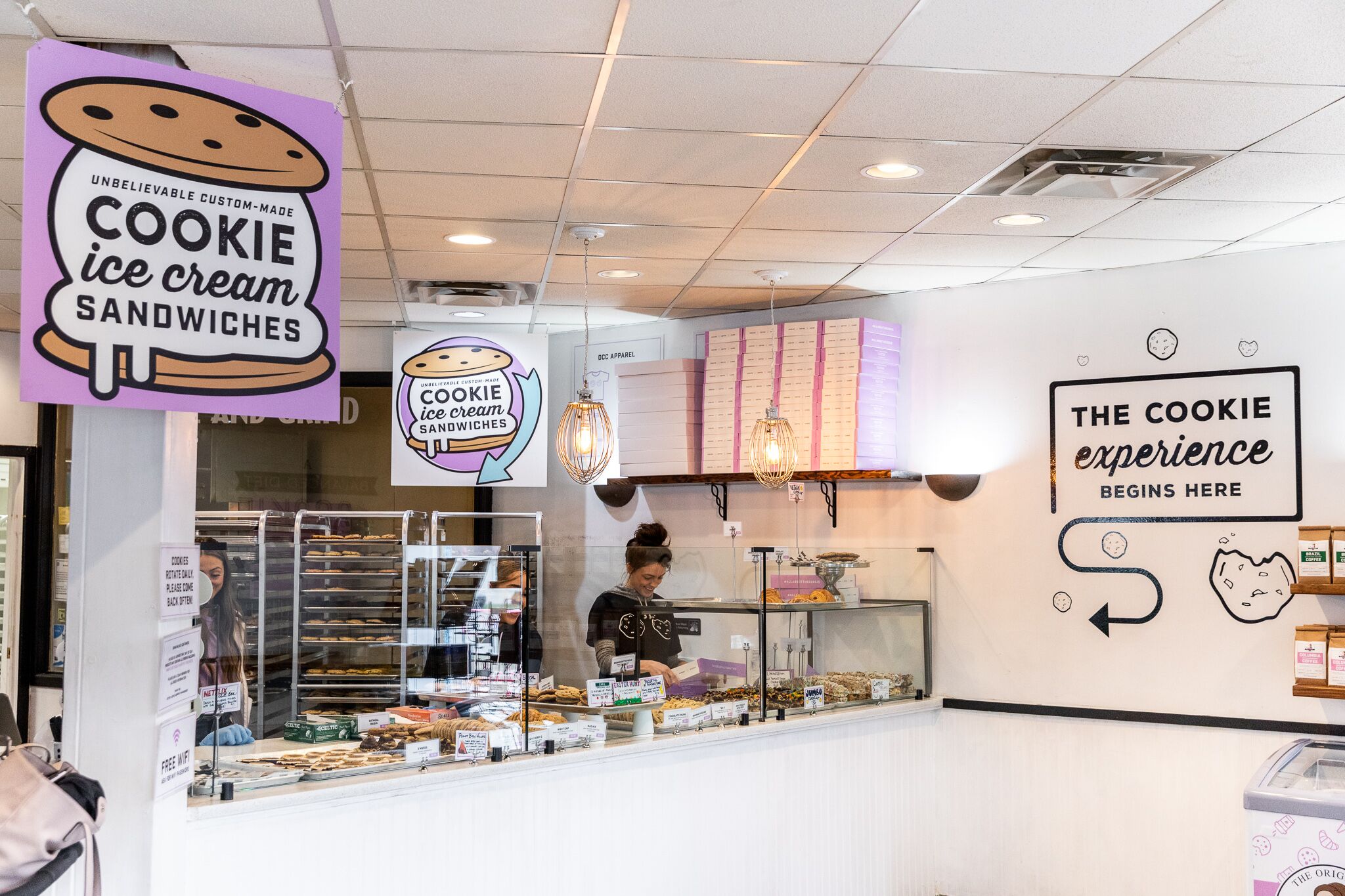 The inside of the Detroit Cookie Co. cafe features white walls and a counter with cookies on trays. Pink signs surround the counter advertising items such as “Unbelievable custom-made ice cream sandwiches.”