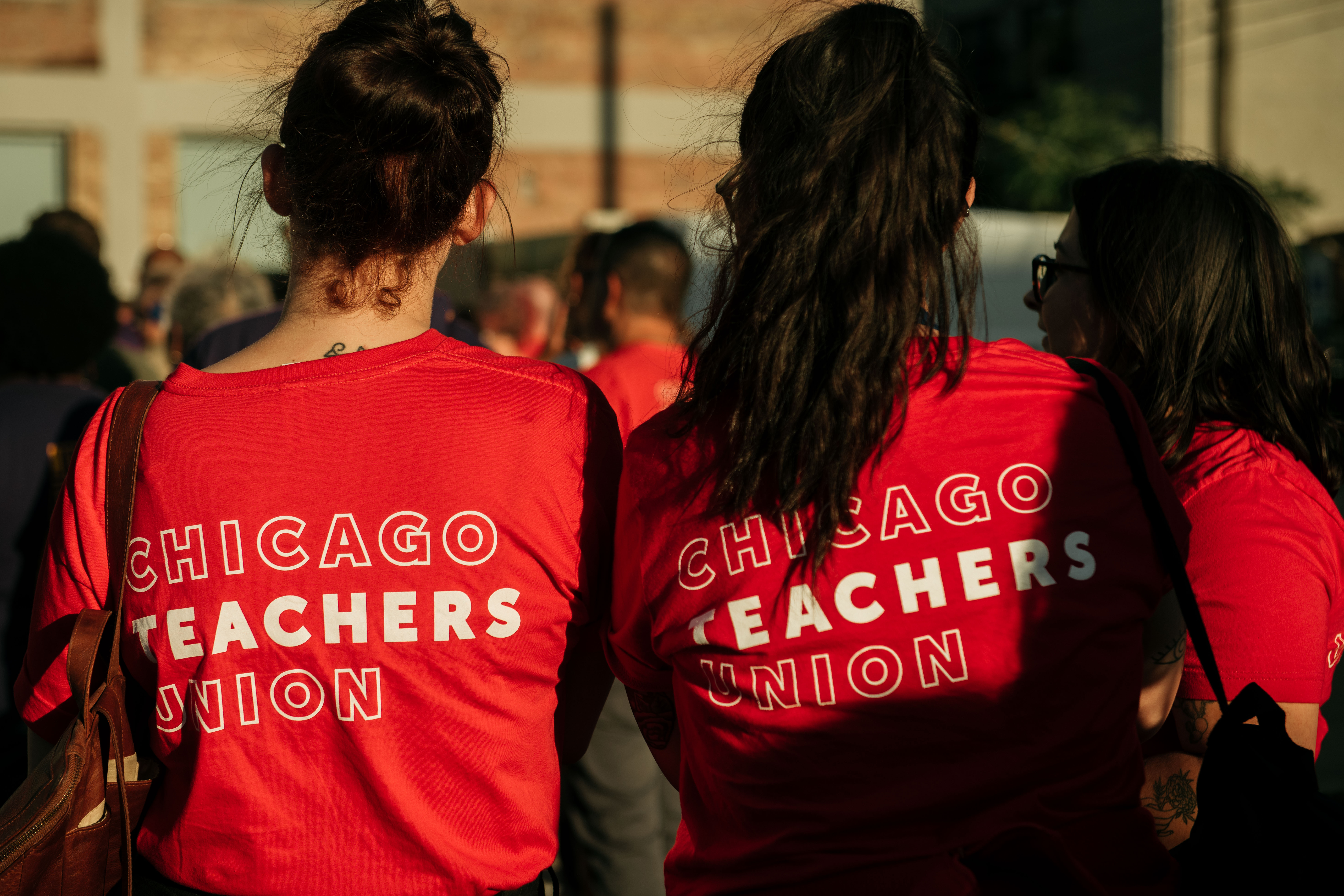 Two women stand together in red shirts with their backs facing. Text on their shirt reads: Chicago Teachers Union.