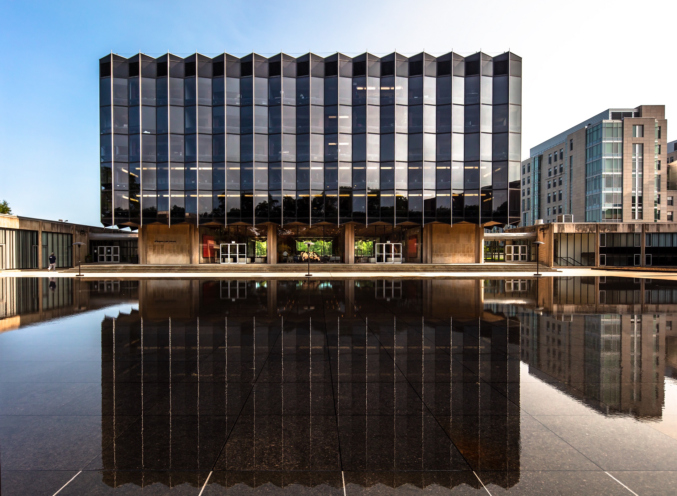 A dark modern building with an angular facade stands before a smooth reflecting pool. 