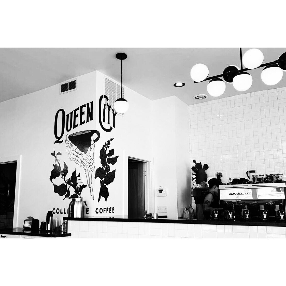 A view of the espresso bar at Queen City Collective Coffee in Five Points with a large graphic showing a tattoo holding a coffee mug on the wall to the left 