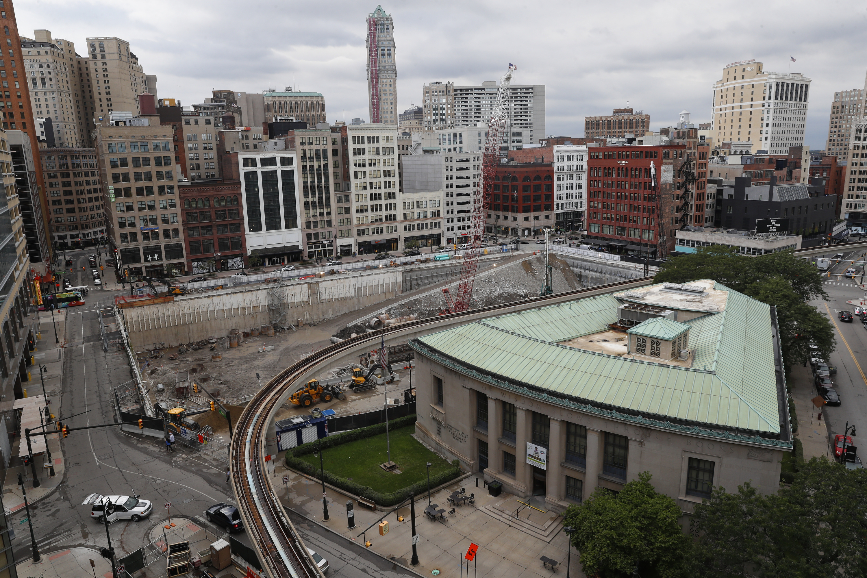 A high view of a rectangular construction site where a foundation is being built below ground. In the foreground is a triangular-shaped building with a copper roof. The background is filled with tall buildings.