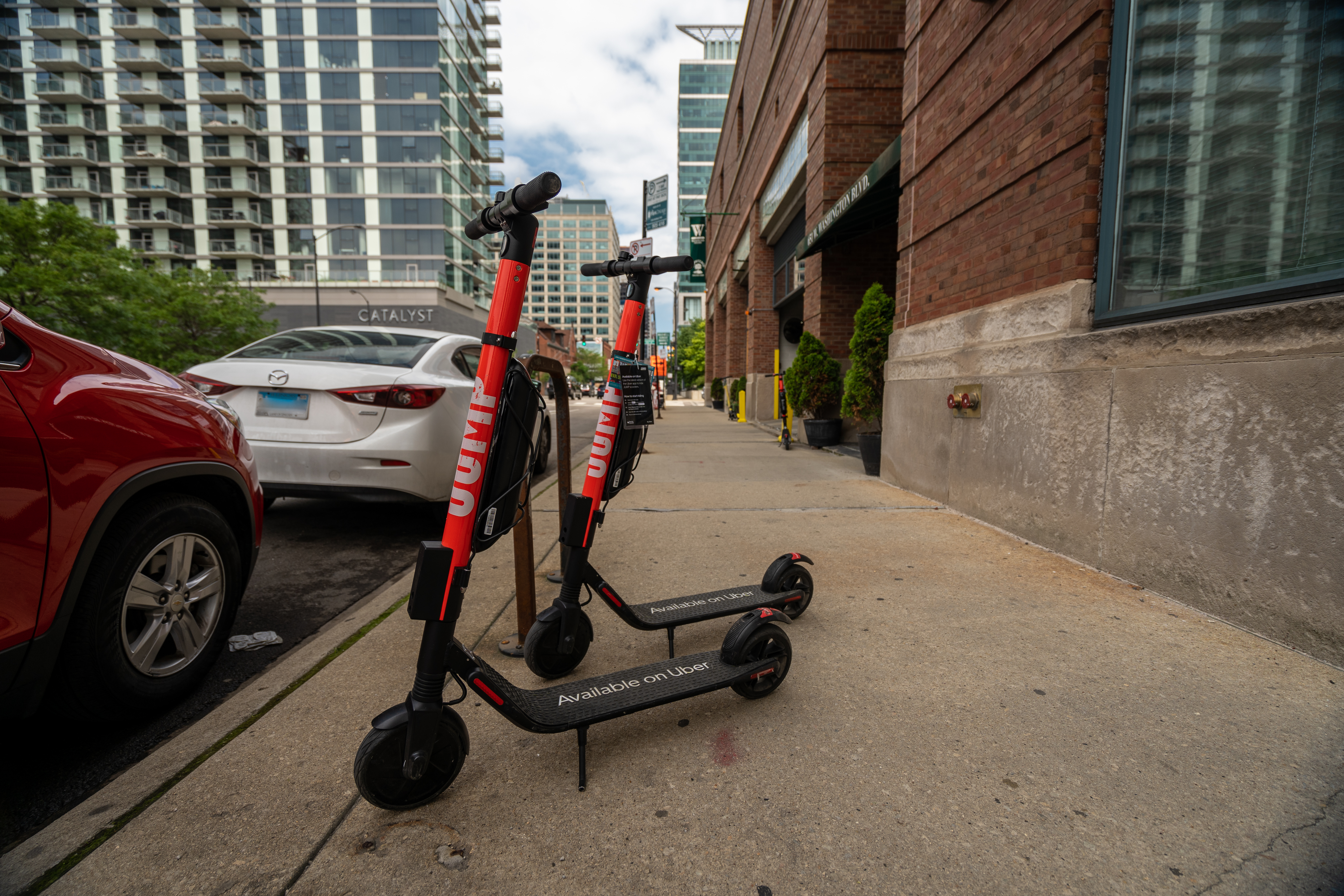 Two scooters parked on a sidewalk next to a brick building and two parked cars. 