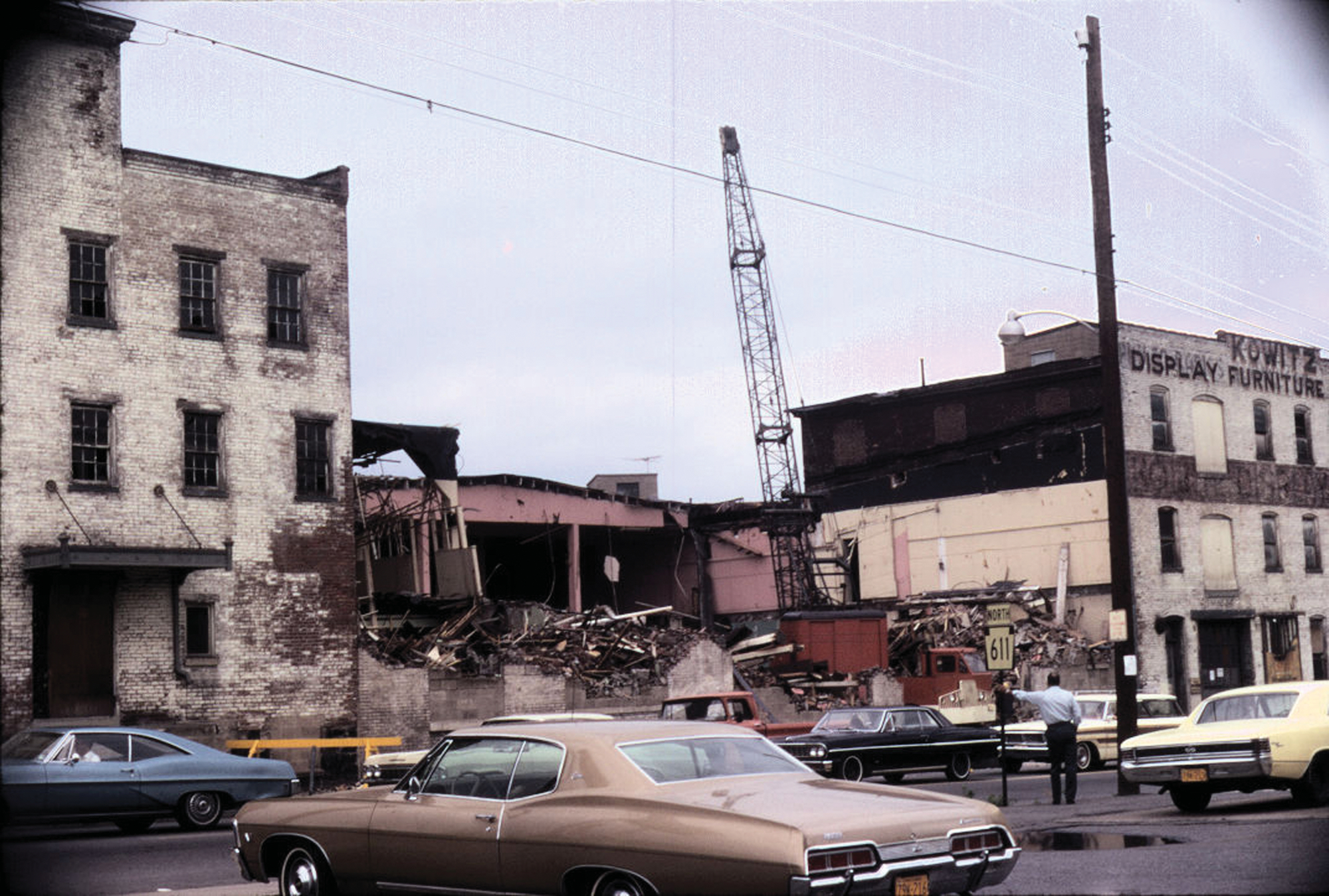A 1970s photograph of an old brick building on a city block under demolition with a man standing by a street sign and cars passing by.