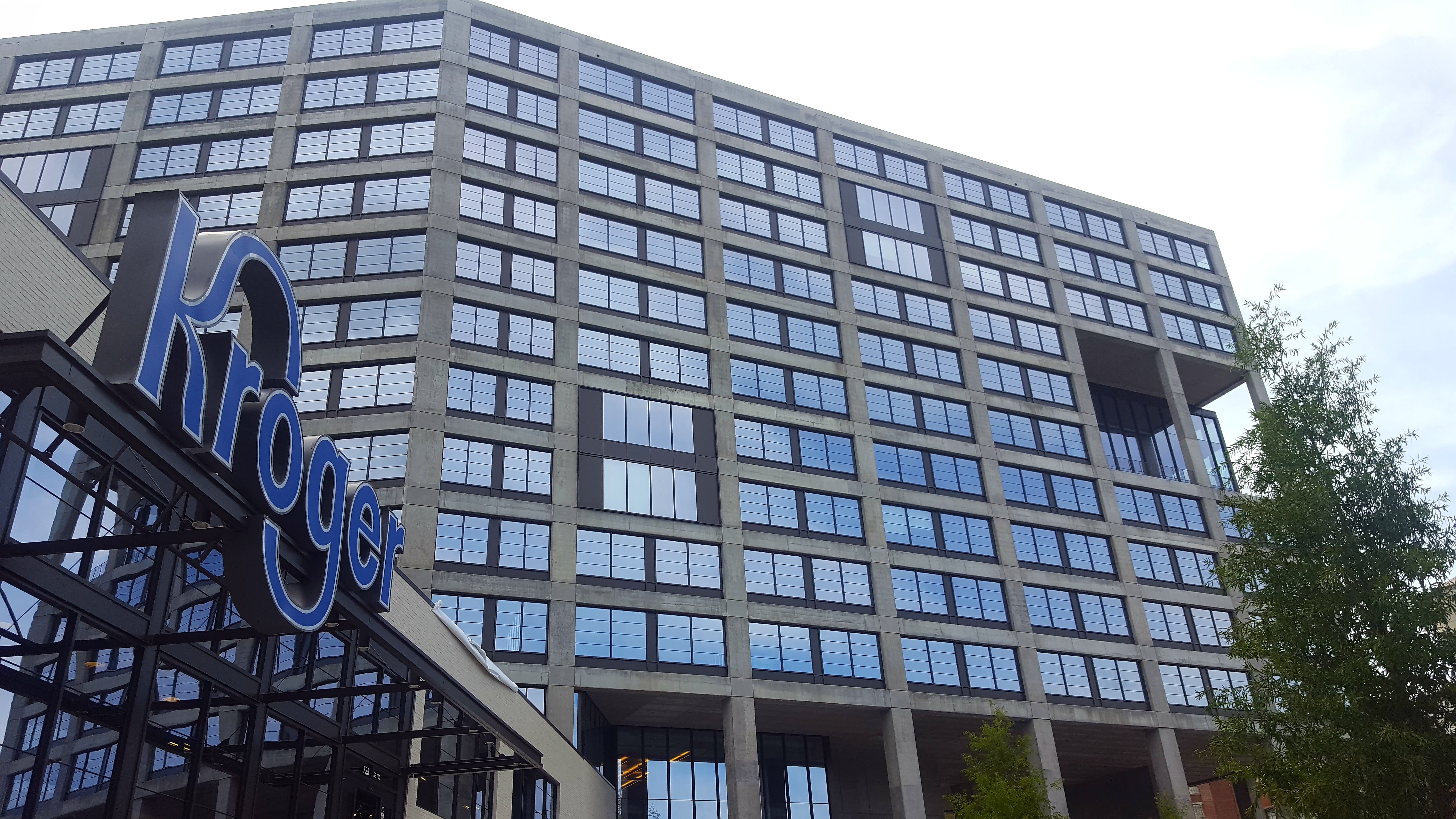 The blue Kroger logo hangs on a two-story building beneath a large, glassy office building.