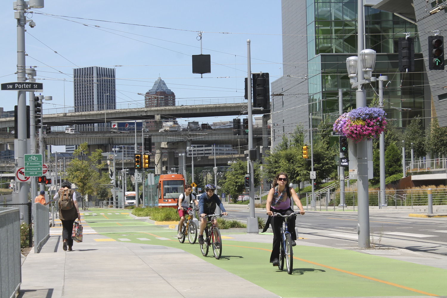 A city street with cyclists riding in bike lanes painted in green, pedestrians walking on a wide sidewalk, and a light rail line.