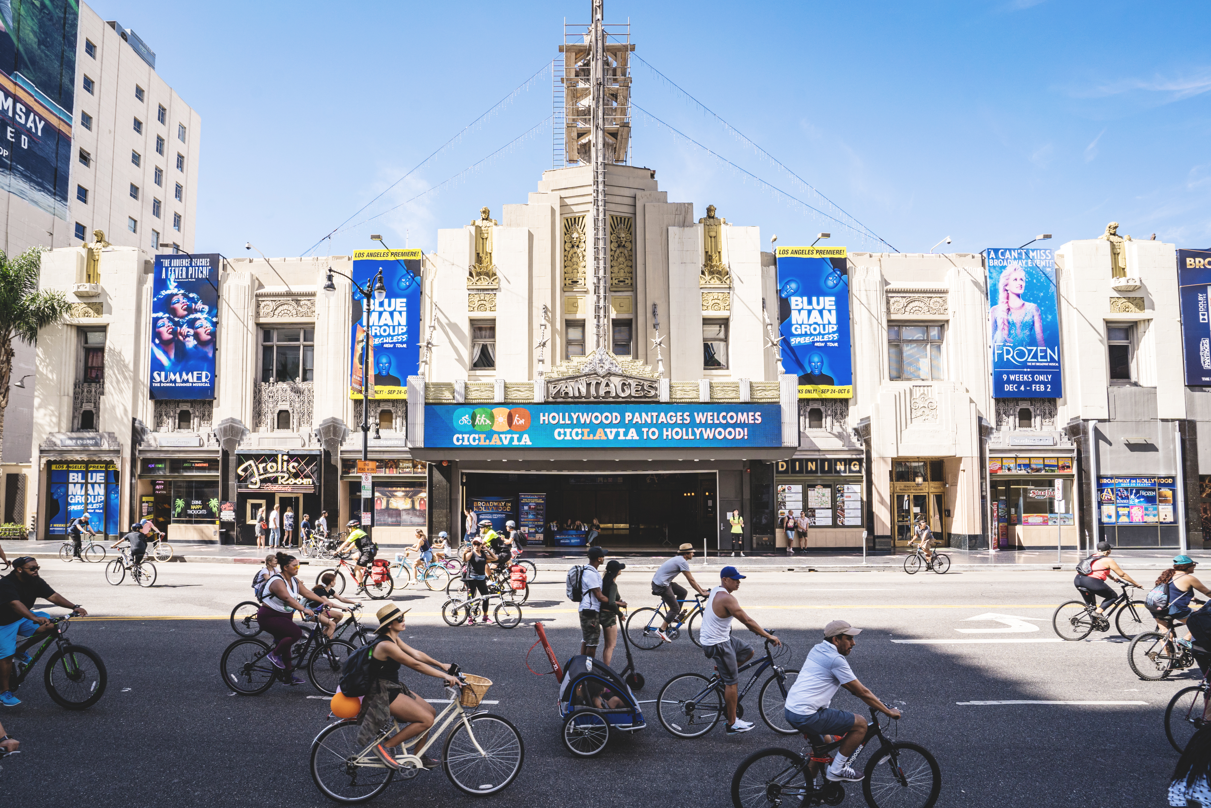 People ride bikes and walk in the street in front of an ornate theater as part of an open streets event in Hollywood.