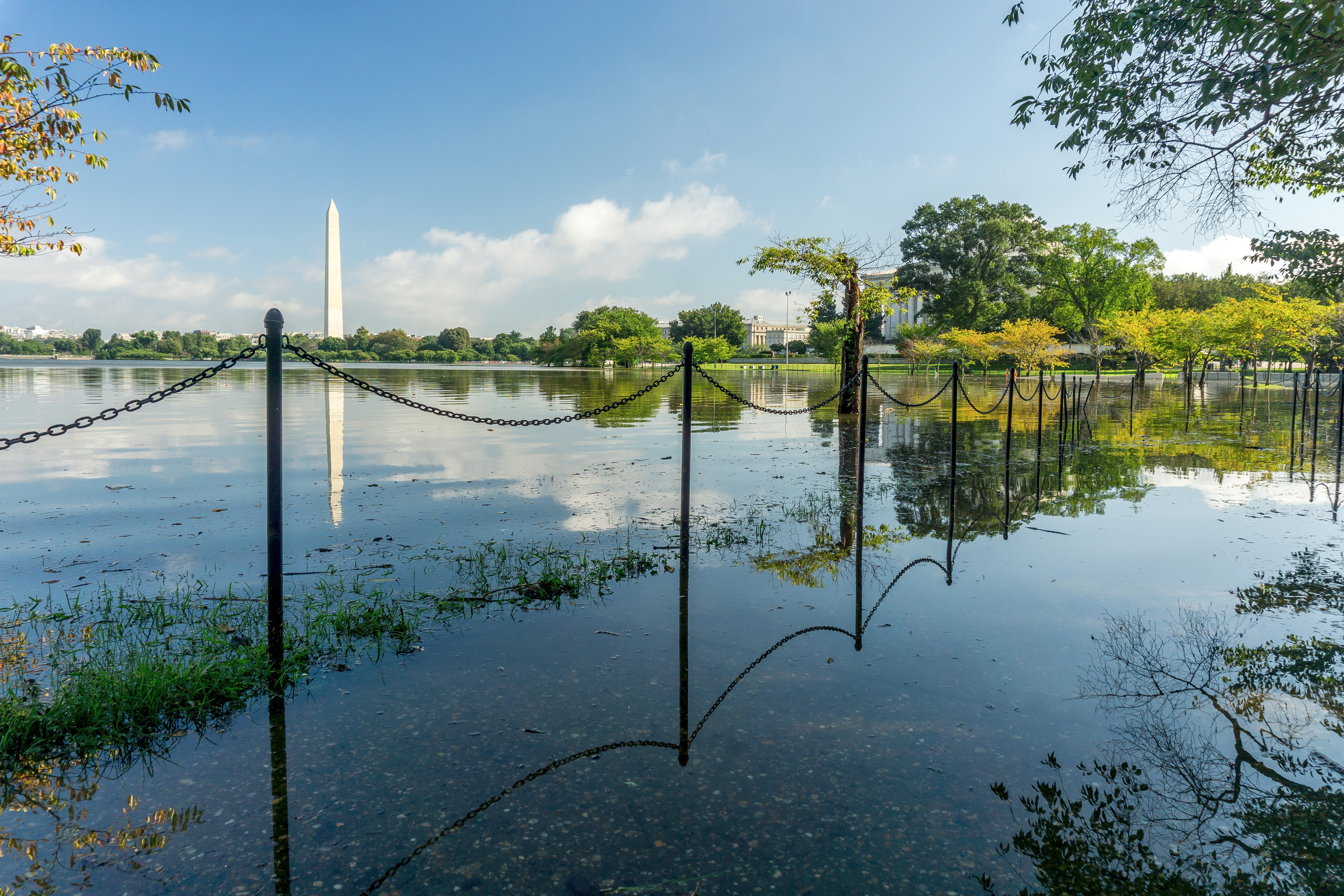 A walkway next to a basin is flooded with a few inches of water. A tall obelisk stands in the background against bright blue skies.
